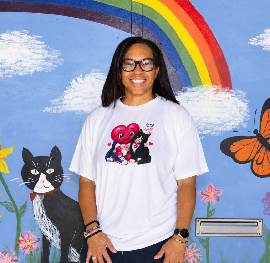 A smiling teacher with glasses and a white T-shirt standing in front of a colorful mural featuring a rainbow, clouds, flowers, a cat, and ladybugs.