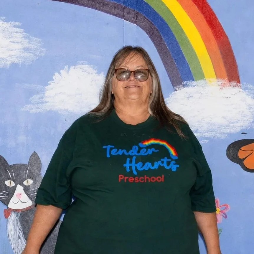 A teacher with glasses and long hair taking a selfie in front of a colorful rainbow mural painted on the wall. The mural includes a green tree with red apples and a blue sky.