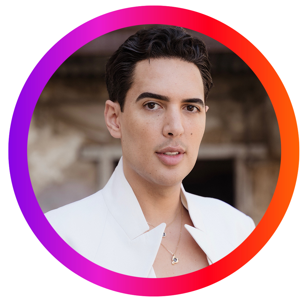 Joven hombre con cabello oscuro y piel clara usando ropa blanca y collar, en un fondo borroso.