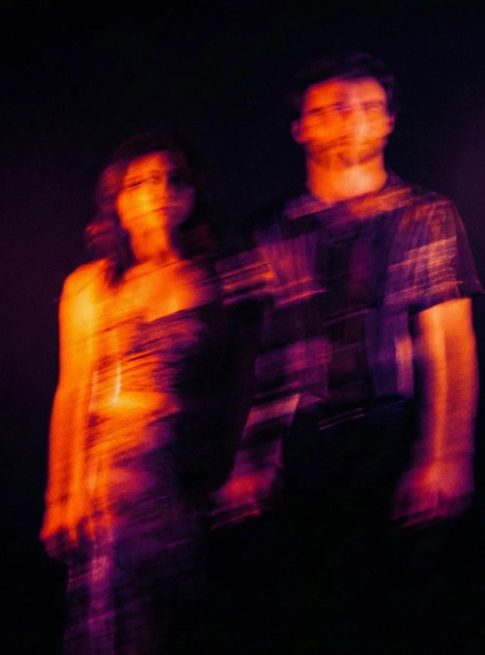 Dos personas, un hombre y una mujer, en un fondo oscuro con iluminación morada y naranja, con efectos de movimiento y desenfoque, creando una imagen artística y abstracta.