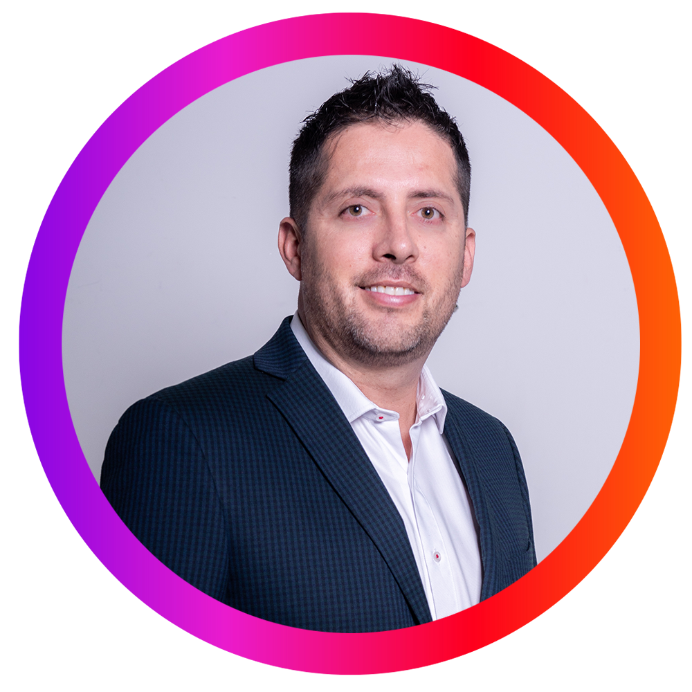 Retrato de un hombre con cabello oscuro, barba y bigote, usando saco azul y camisa blanca, contra fondo gris con marco de colores rosa, morado y naranja.