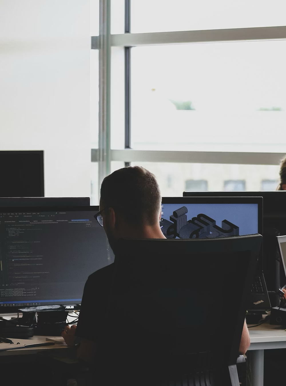 Persona trabajando en una oficina con computadoras, vista de espaldas.