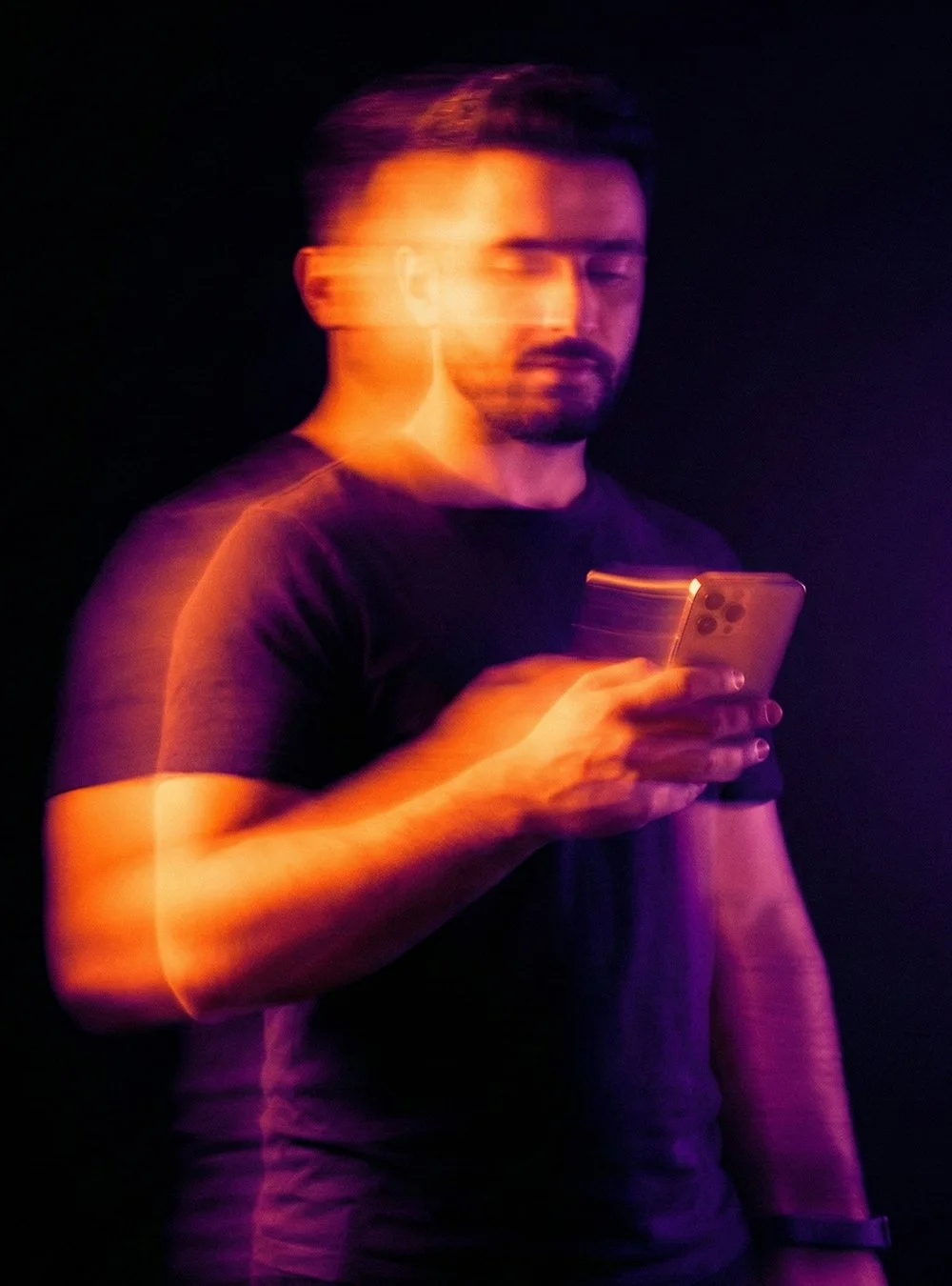 Hombre con barba y camiseta negra viendo su teléfono móvil, efecto de movimiento y luces de colores en un fondo negro.