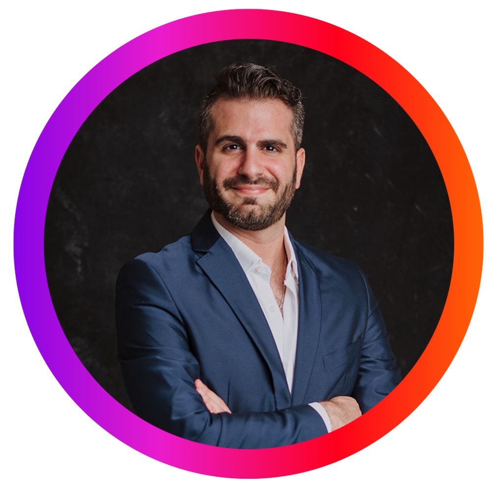 Hombre con barba, vestido con saco azul y camisa blanca, sonriendo con brazos cruzados, fondo oscuro, rodeado por un marco circular de color degradado en tonos rosas, morados y naranjas.