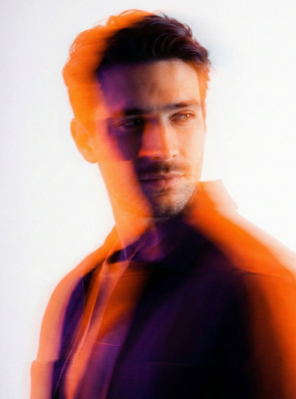 Retrato artístico de un hombre con cabello oscuro y barba, con efectos de doble exposición en tonos naranja y morado, luciendo una chaqueta negra y fondo blanco.