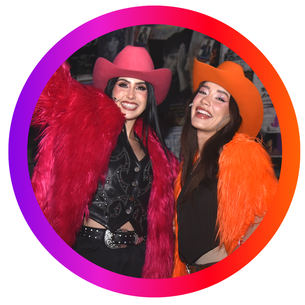 Dos mujeres vestidas con sombreros y chaquetas de peluche en colores rosa y naranja, sonriendo en un fondo oscuro con decoraciones.