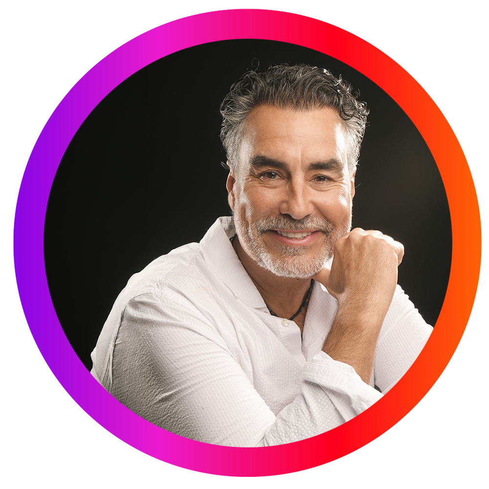 Retrato de un hombre de cabello gris y barba, sonriendo, con fondo negro, rodeado por un marco circular en tonos morados, rosados y rojos.