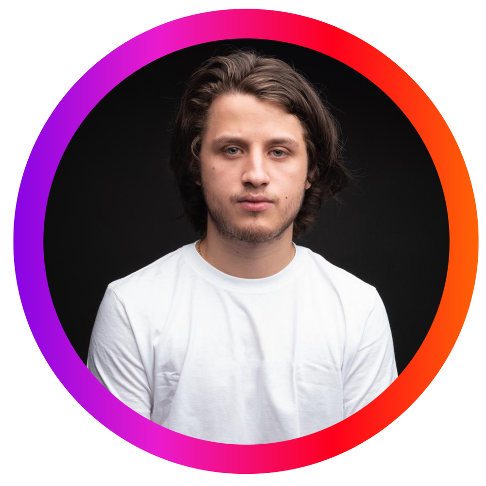 Un joven con cabello largo y rizado, de piel clara, viste una camiseta blanca, posa frente a un fondo negro.