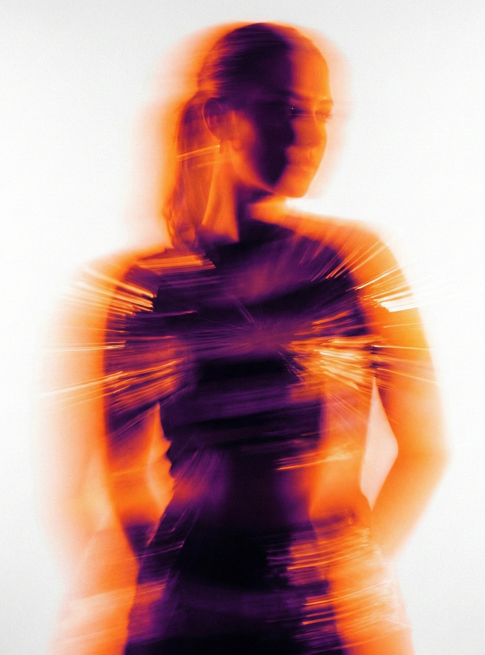 Mujer con apariencia de movimiento o distorsión, vestida con ropa oscura, en tonos anaranjados y púrpuras, con fondo claro.