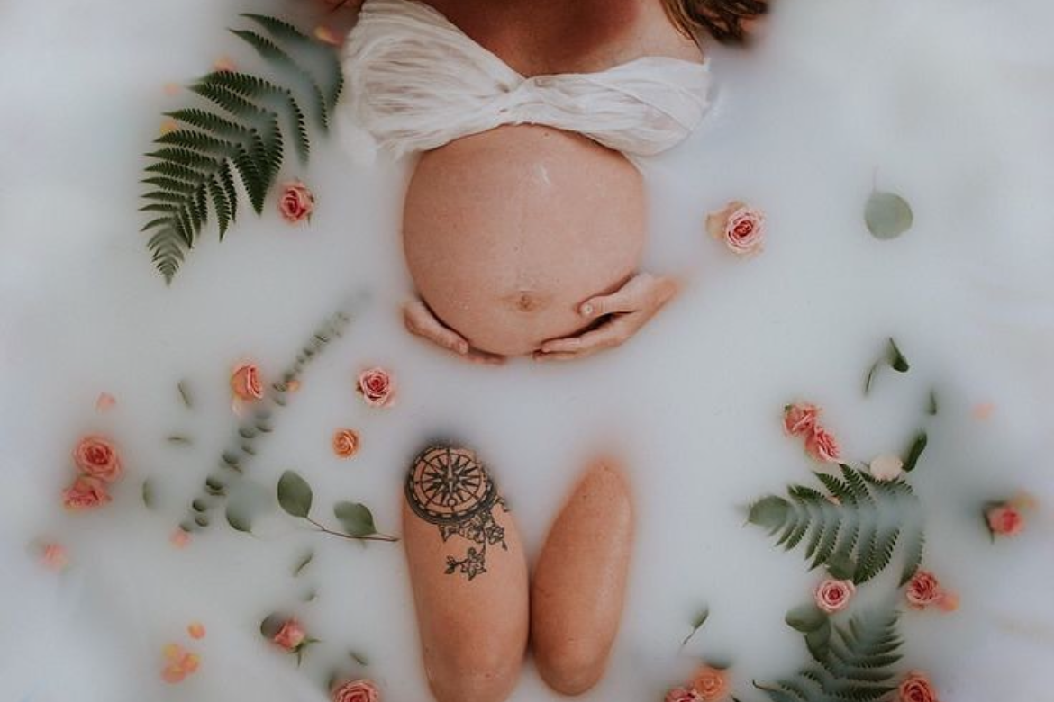 Maternità in un bagno di latte, con il ventre di una donna nuda coperto da un tessuto bianco, circondata da fiori rosa, foglie verdi, pietre e tatuaggi.