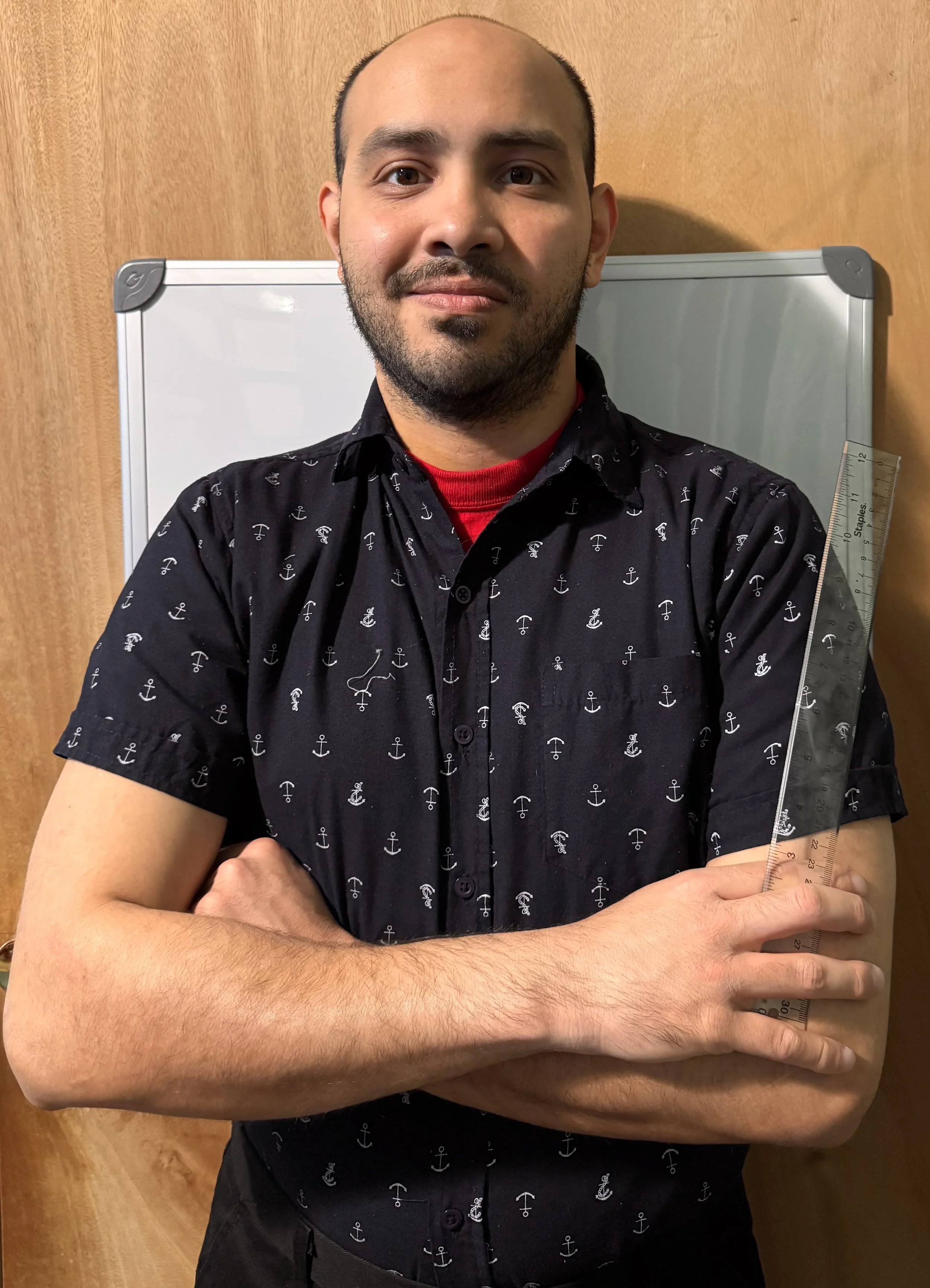 A man with a shaved head and beard, wearing a black shirt with small white anchors, standing in front of a wooden door and whiteboard, holding a ruler across his chest with arms crossed.
