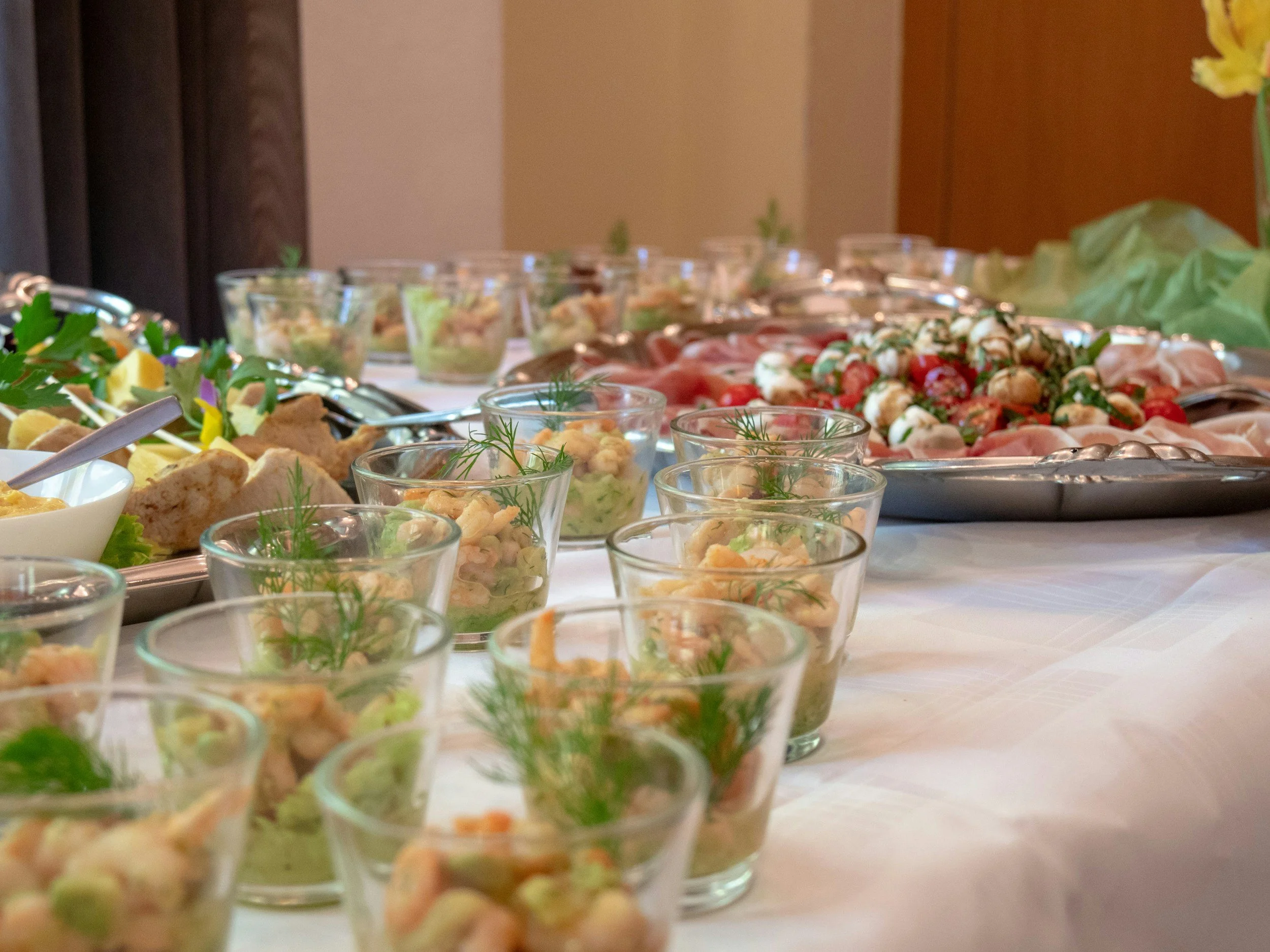 A table of assorted appetizers including small glasses of seafood salad garnished with dill, vegetable salads, and a tray of cherry tomatoes with mozzarella and basil, set up on a white tablecloth for a party or gathering from Benny's Catering.