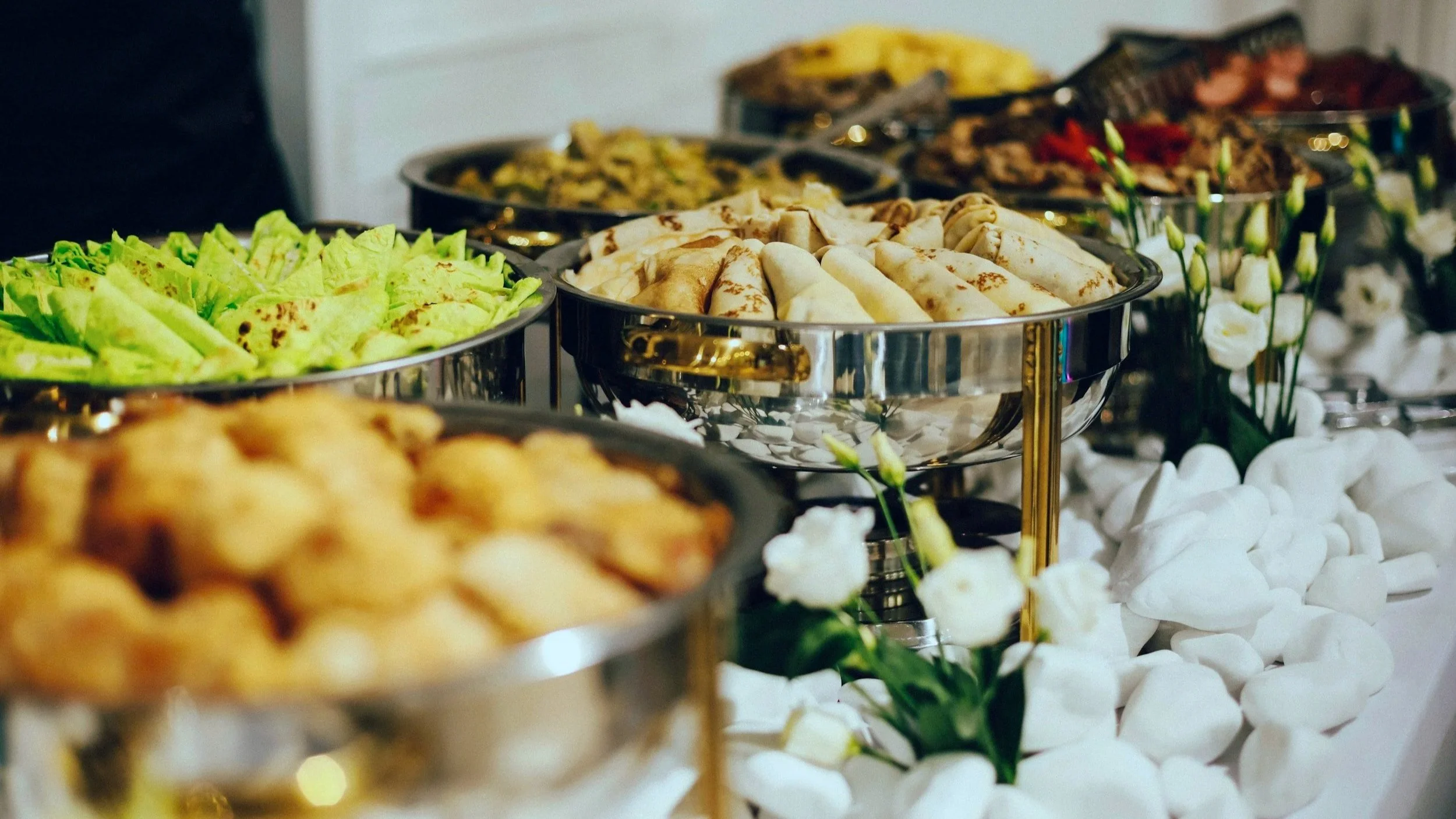A buffet table with various dishes, including plate of lettuce wraps, grilled vegetables, and rolled tortillas, decorated with white flowers and stones from Benny's Catering in Greeley