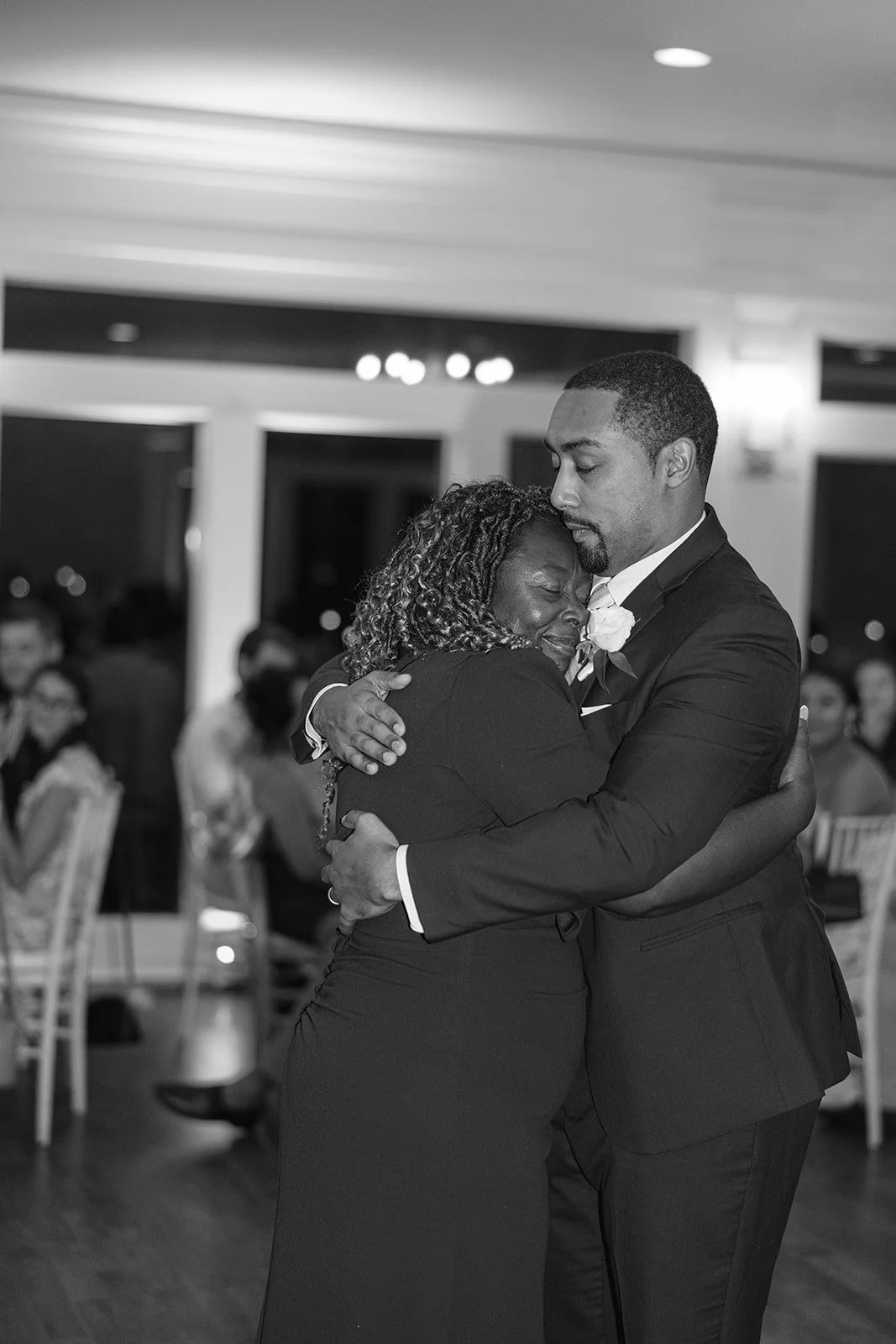 A black and white photo of a couple hugging each other at a wedding reception, with seated guests in the background.