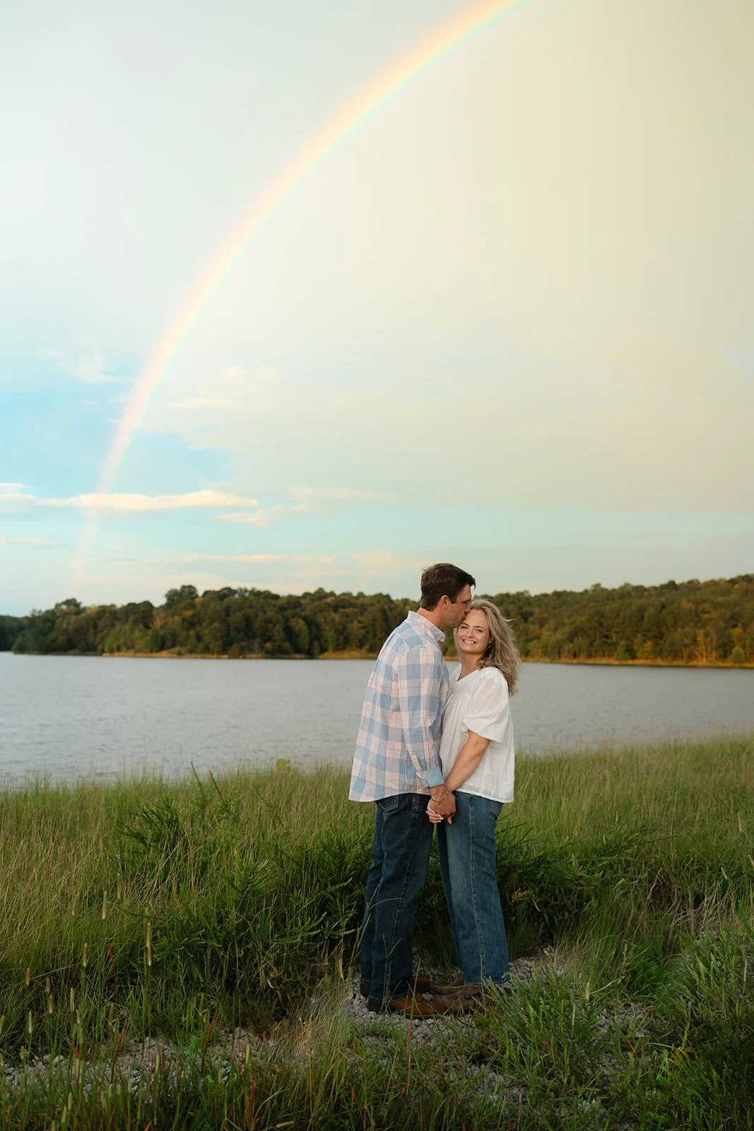 A couple standing on grass near a lake, holding hands and kissing, with a rainbow in the sky above them.