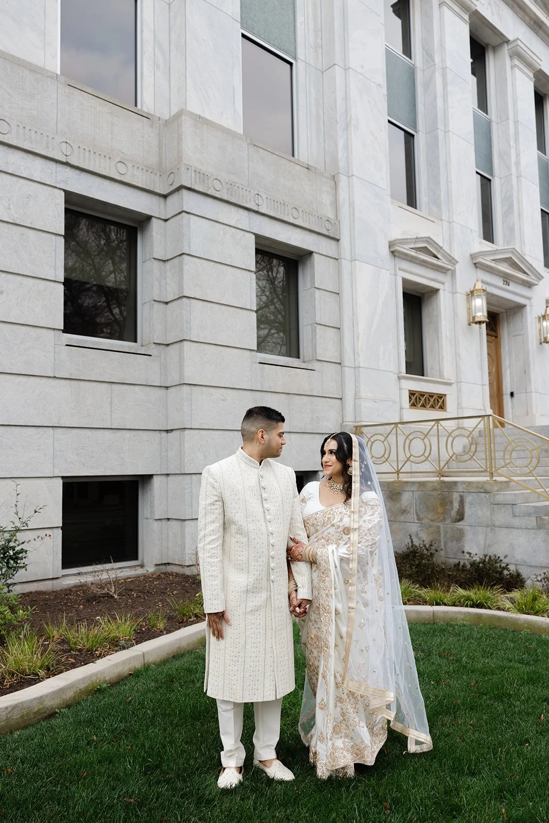 A man and woman dressed in traditional South Asian wedding attire stand on a lawn in front of a white building, holding hands and gazing at each other.