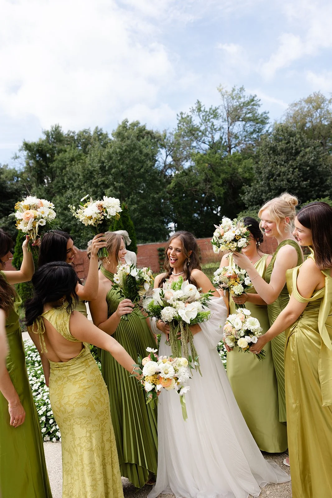 Bride in a white wedding dress and bridesmaids in yellow and green dresses holding bouquets, smiling outdoors.
