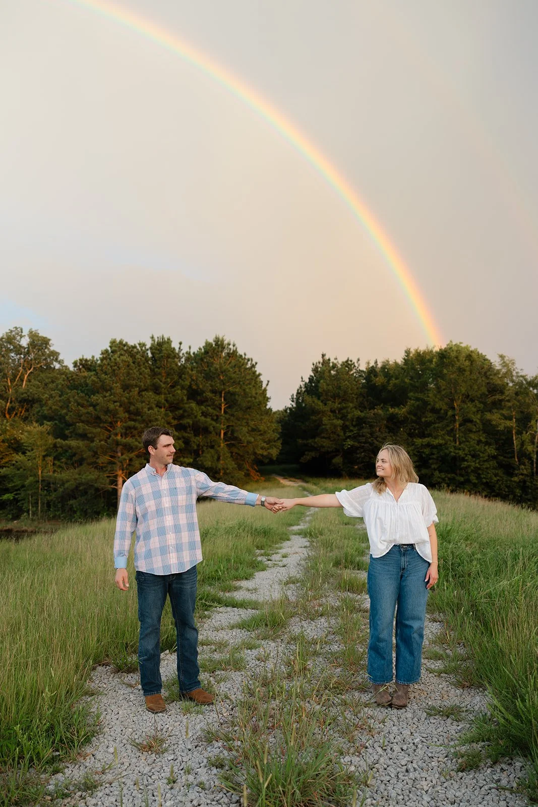 A couple standing on a gravel path in a grassy field, holding hands and smiling at each other under a rainbow in the sky. The background features trees and a cloudy sky.