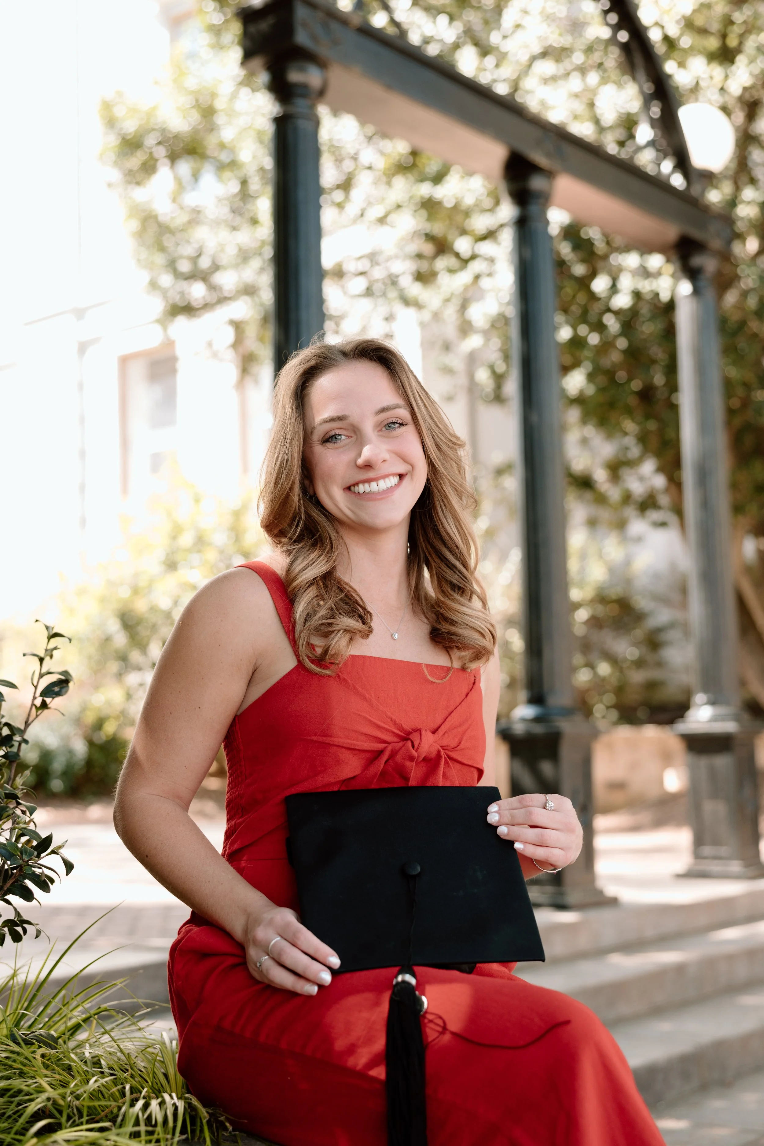 A young woman in a red dress holds a black graduation cap while sitting outdoors on steps, smiling at the camera.