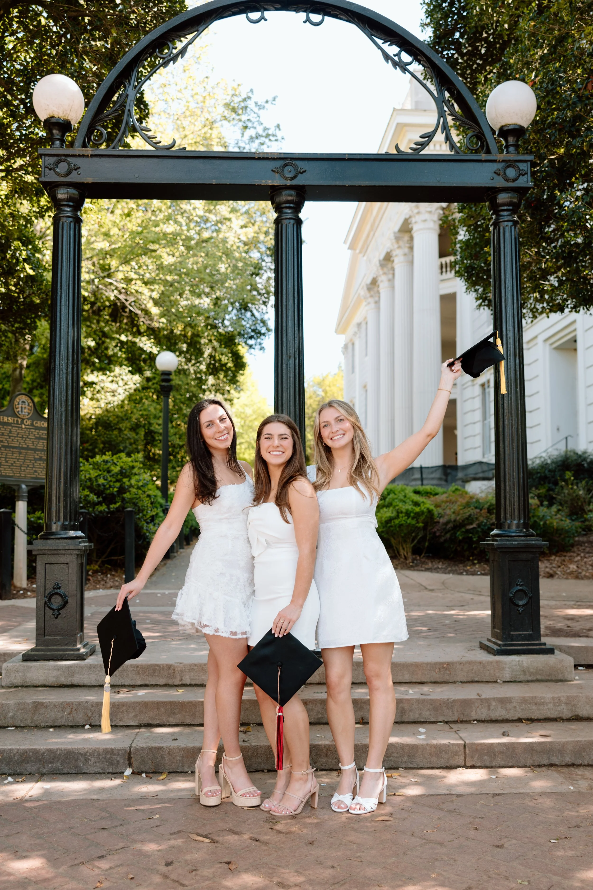 Three young women in white dresses celebrating graduation, holding black caps with tassels, standing on steps in front of a historic building with columns and greenery.