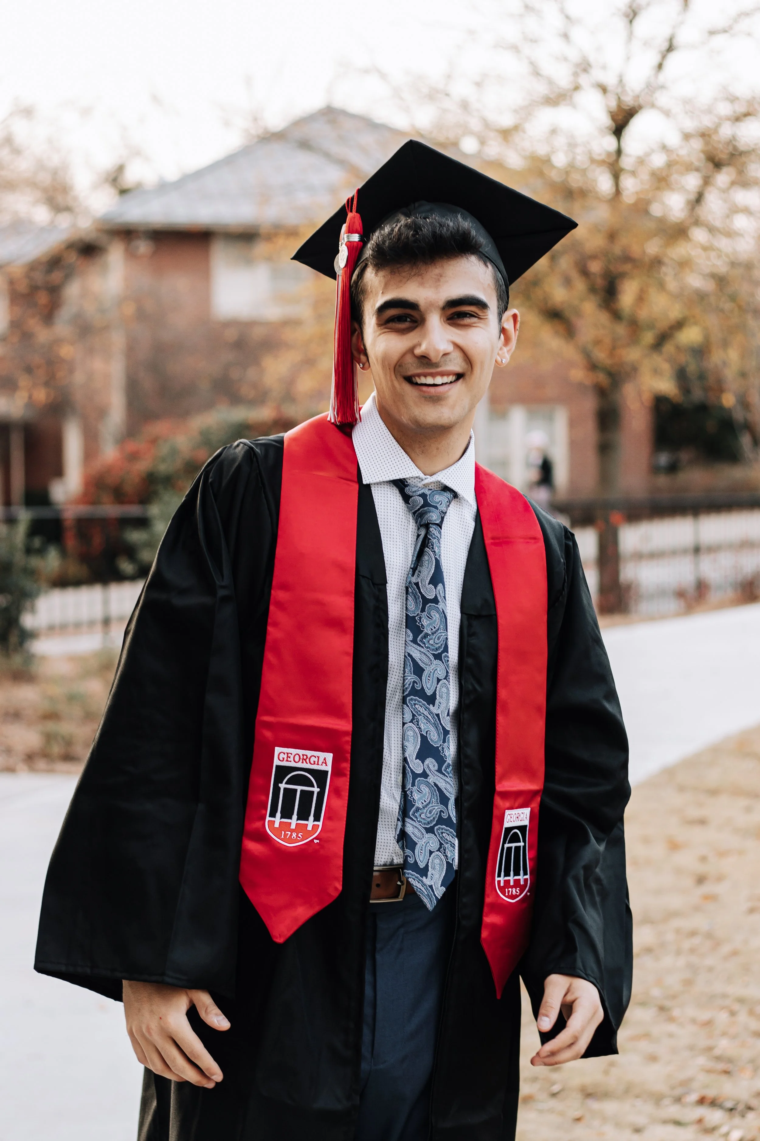 A young man in a graduation gown and cap, smiling outdoors with trees and a house in the background.