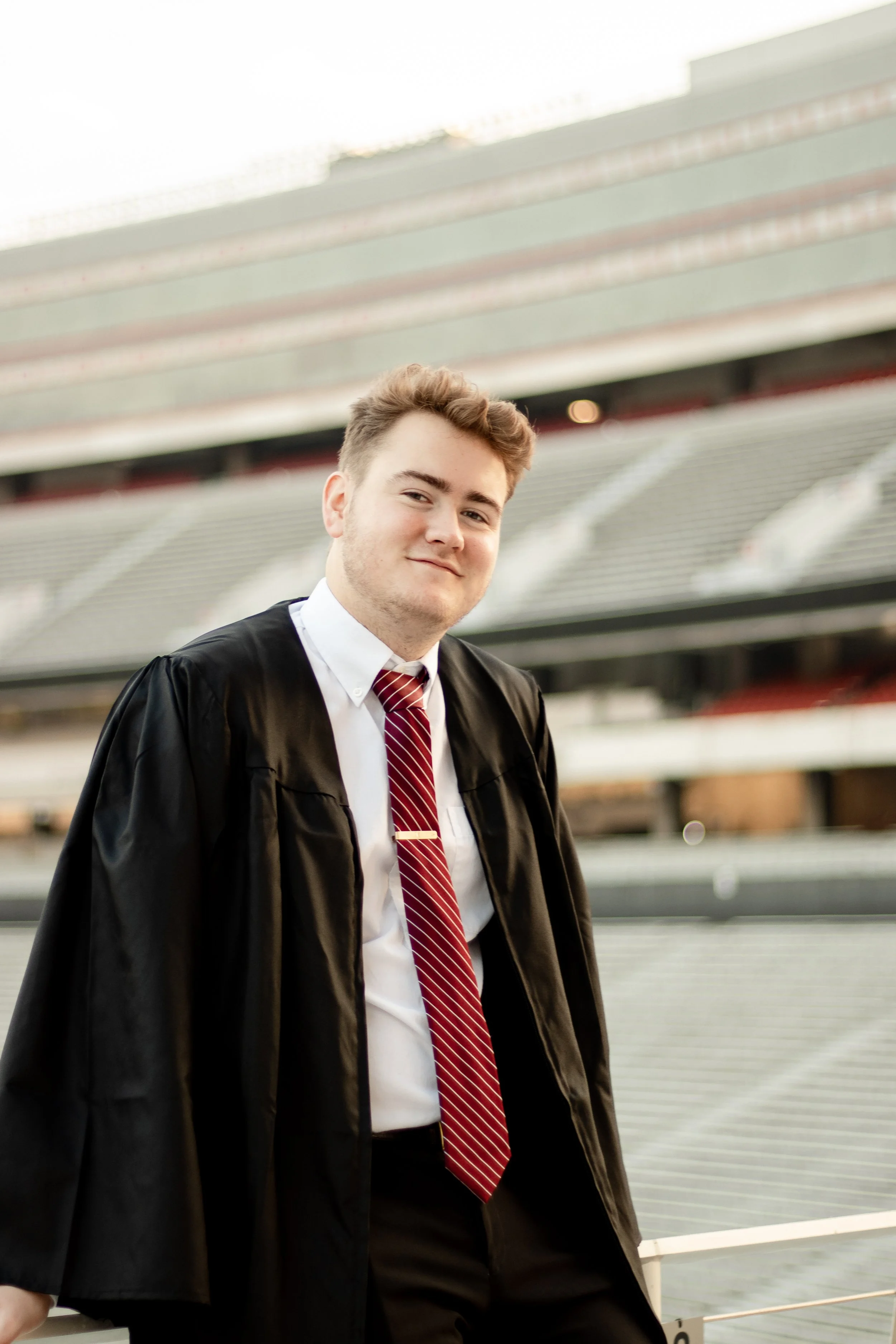 A young man in a graduation gown and tie standing in a stadium.