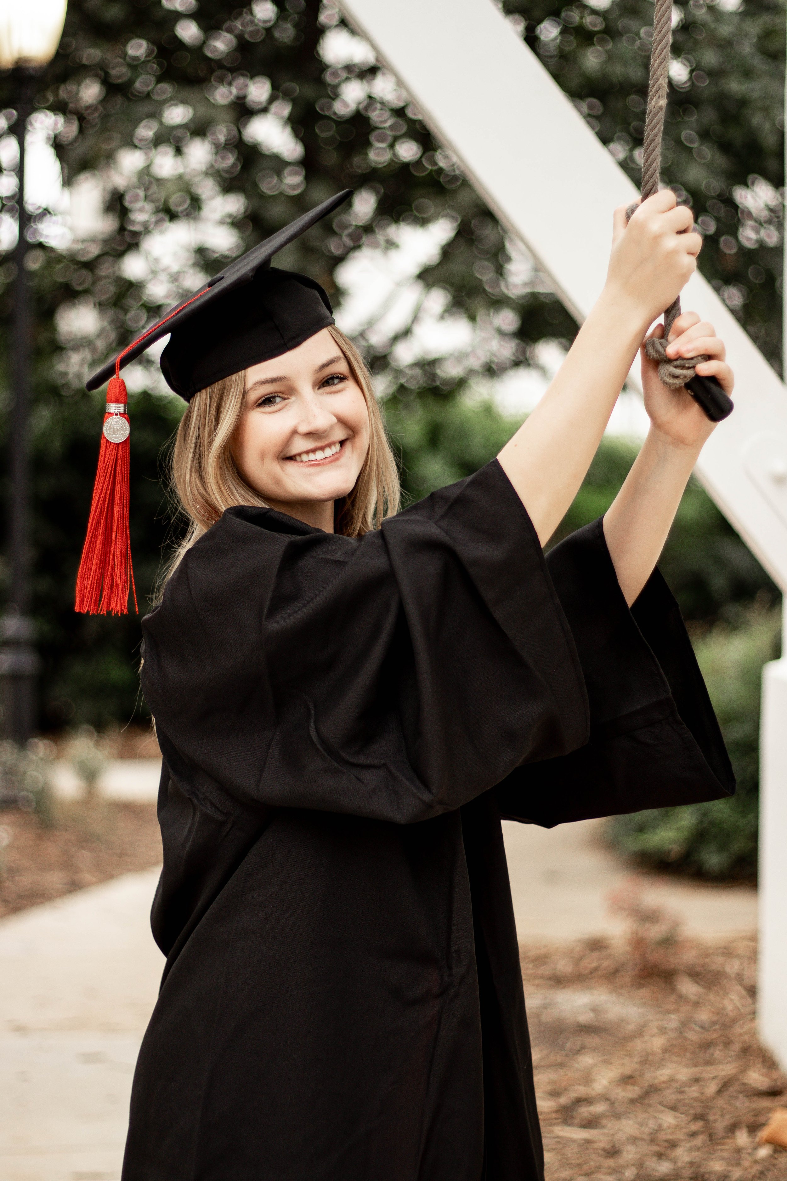 A young woman dressed in a black graduation gown and cap with a red tassel, smiling while holding a rope.