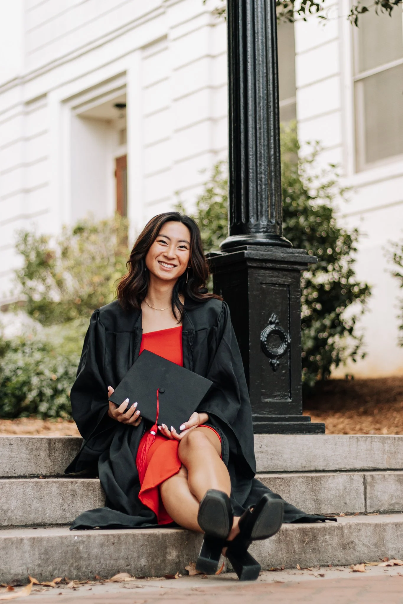 A young woman in a graduation cap and gown sitting on steps outside, smiling and holding her diploma.