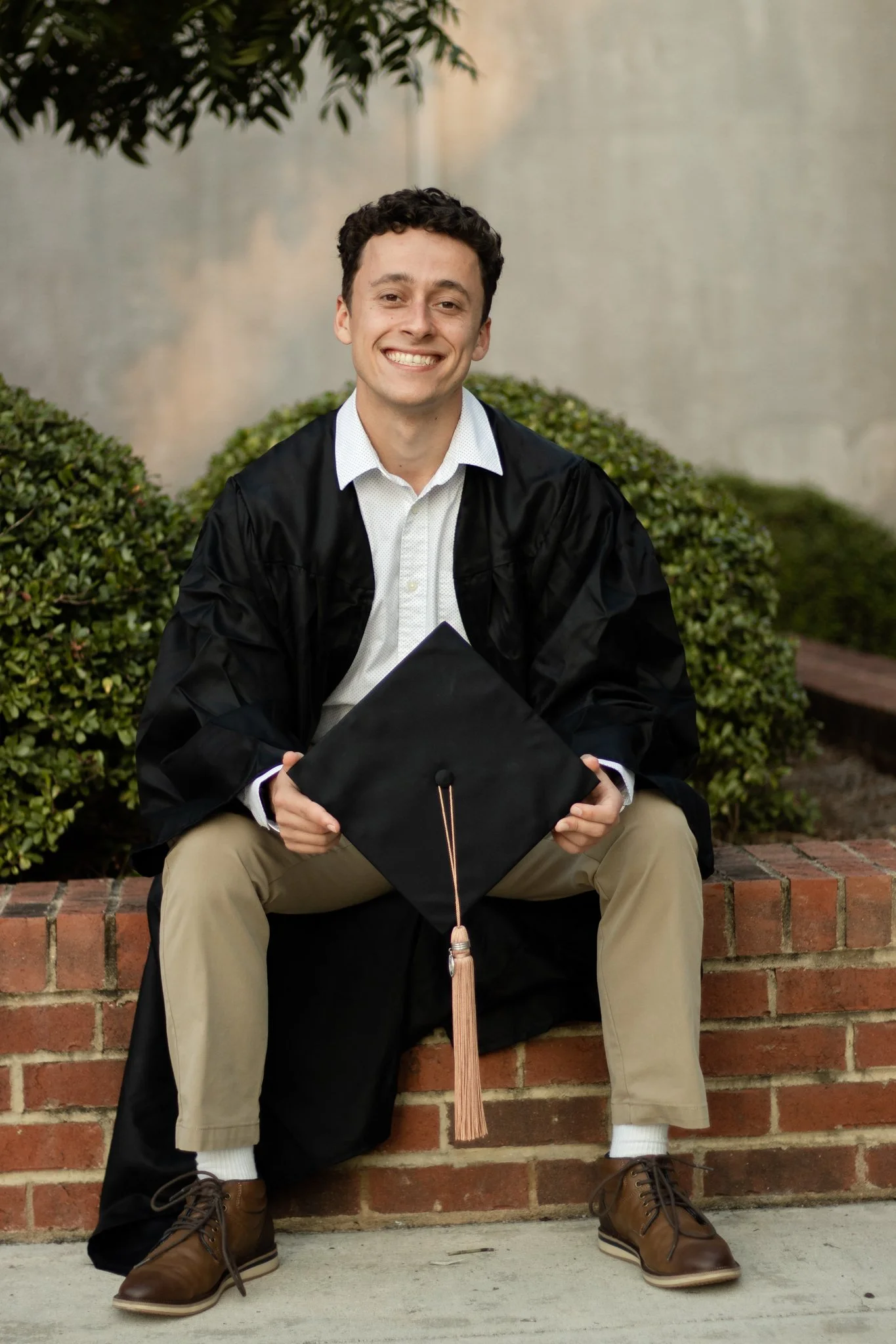 A young man in graduation attire, sitting on a brick ledge outdoors, holding a graduation cap, smiling at the camera.