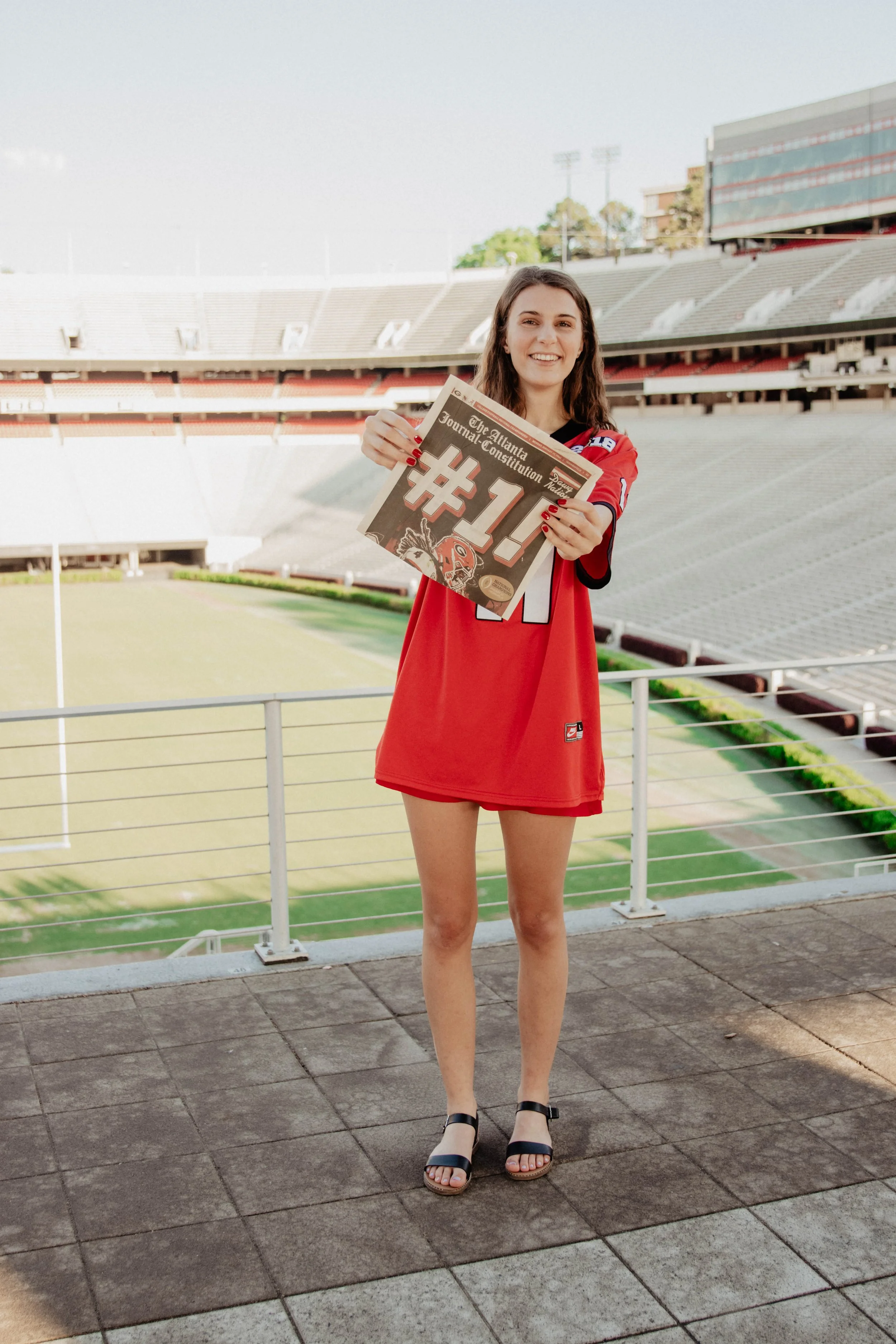A young woman standing on a stadium balcony, holding a newspaper with a large '#1' headline, wearing a red sports jersey, smiling.