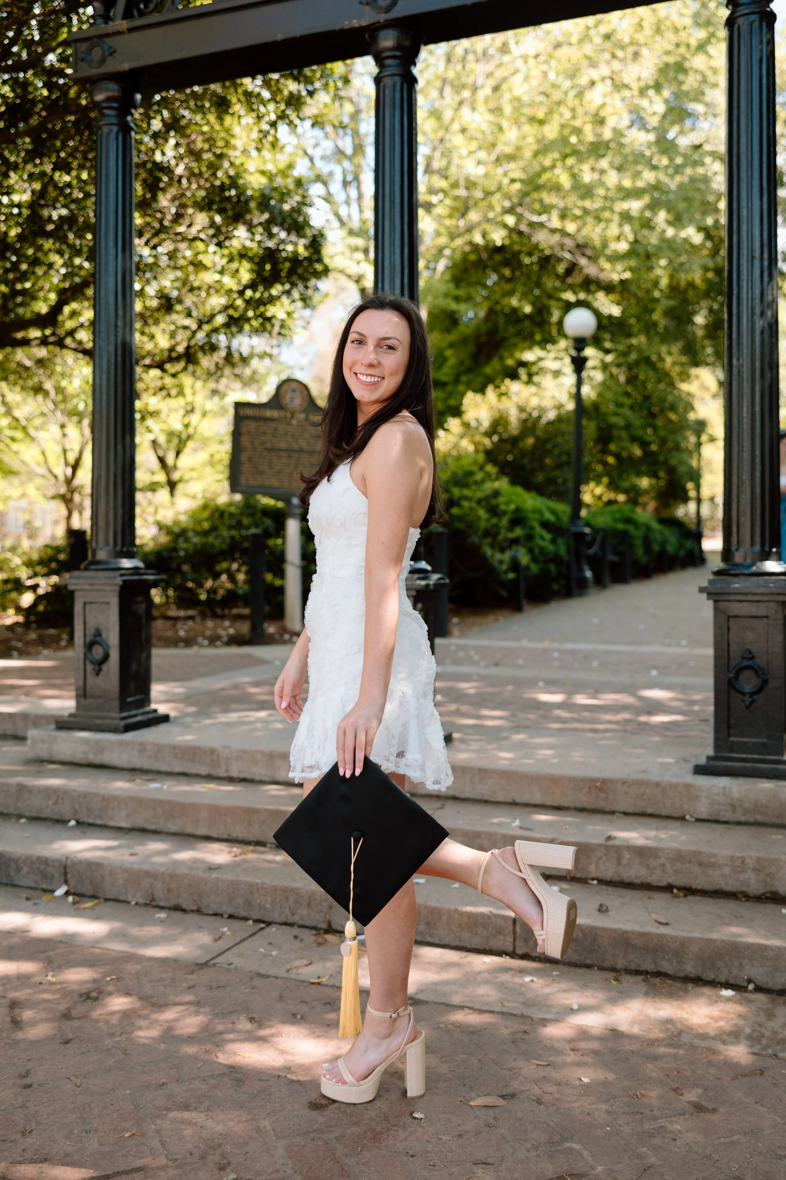 A young woman in a white dress holding a graduation cap and standing on steps in a park.