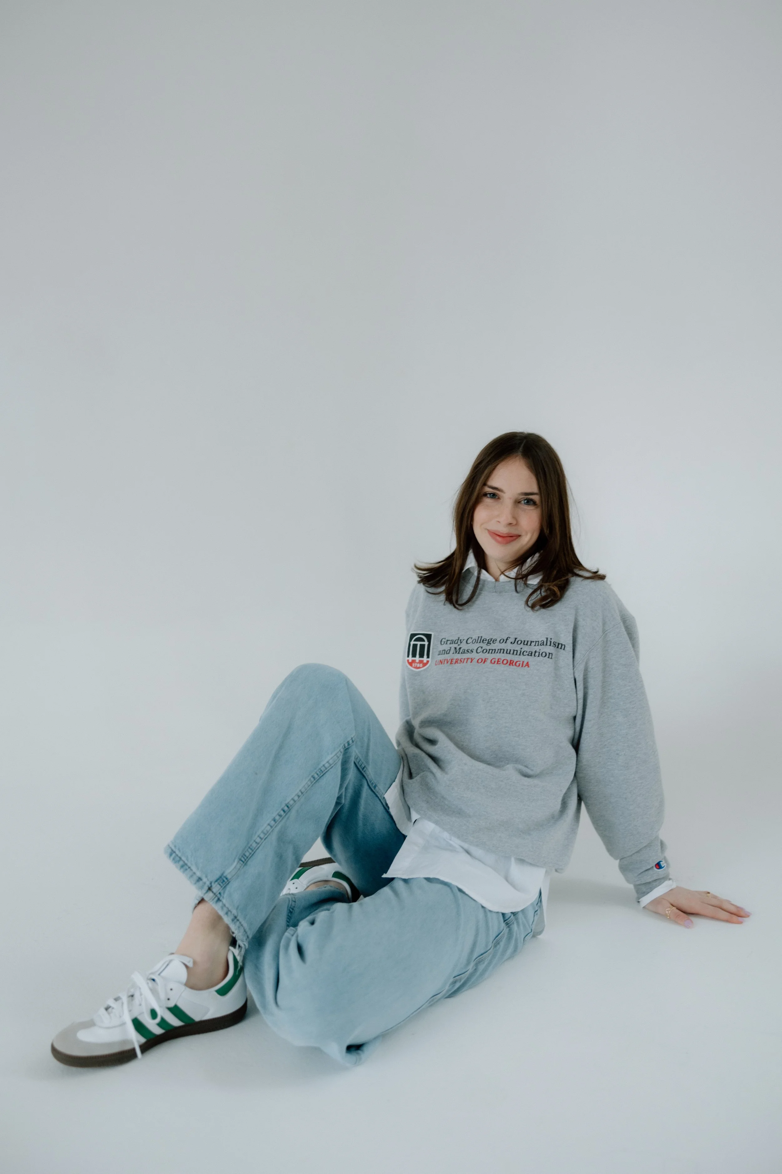 Young woman sitting on the floor, smiling, wearing a gray sweatshirt with university logo and text, light blue jeans, and white sneakers with green stripes, against a plain white background.
