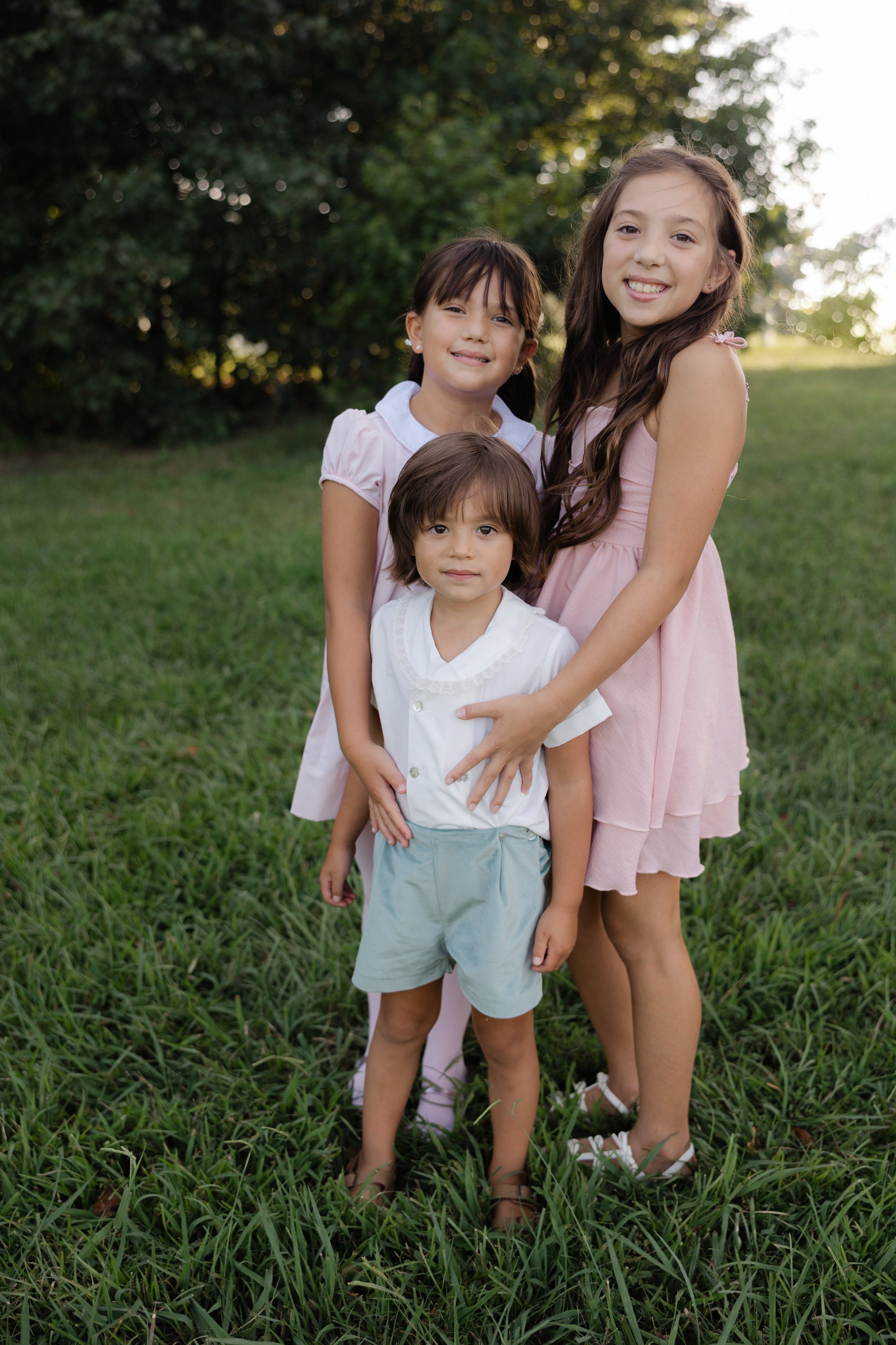 Three children, two girls and a boy, standing outdoors on grass with trees in the background. The girls are wearing pink dresses, and the boy is dressed in a white shirt and light shorts. The children are smiling and looking at the camera.