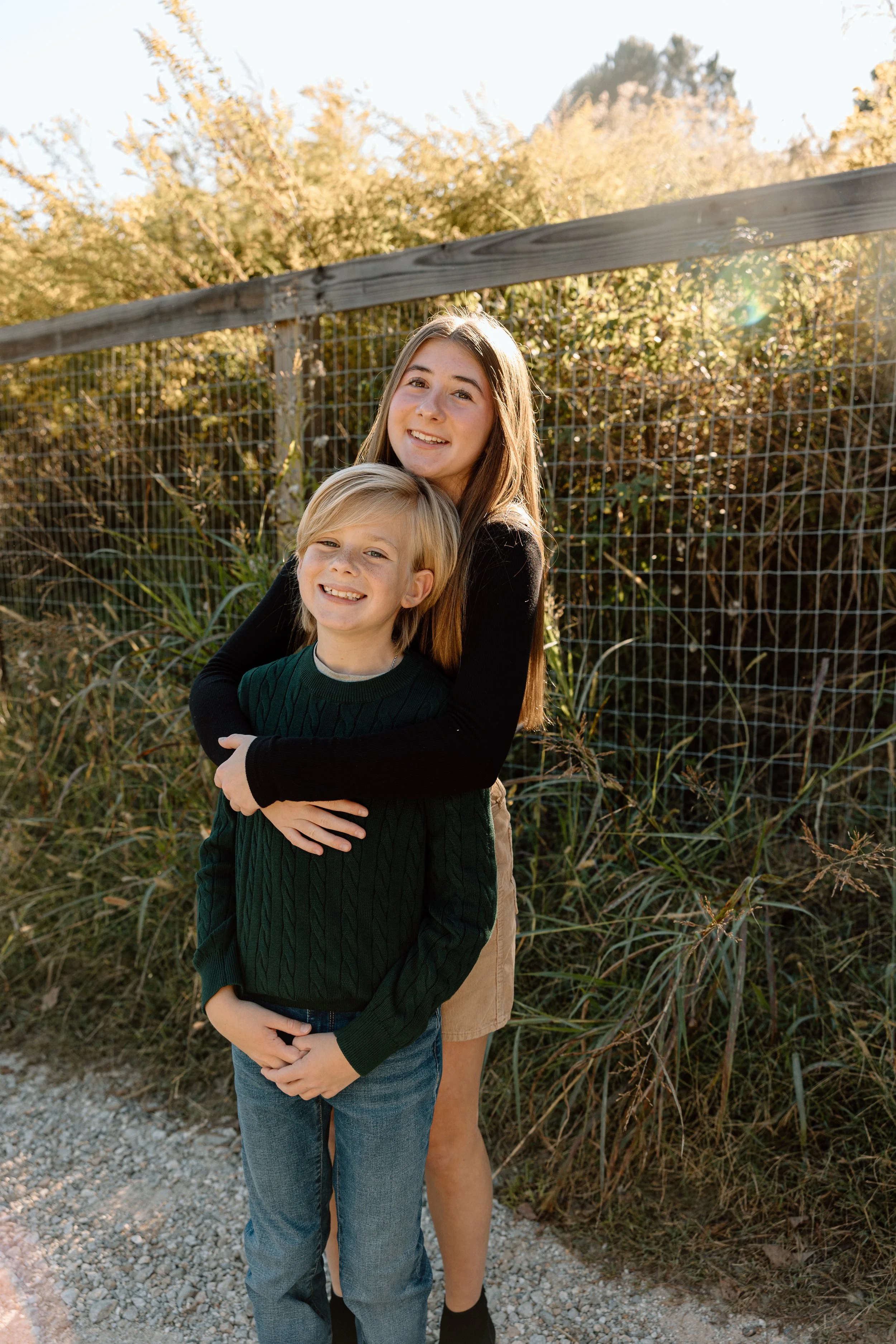 A young woman and a young boy smiling outdoors at sunset, standing in front of a wire fence and tall grass.