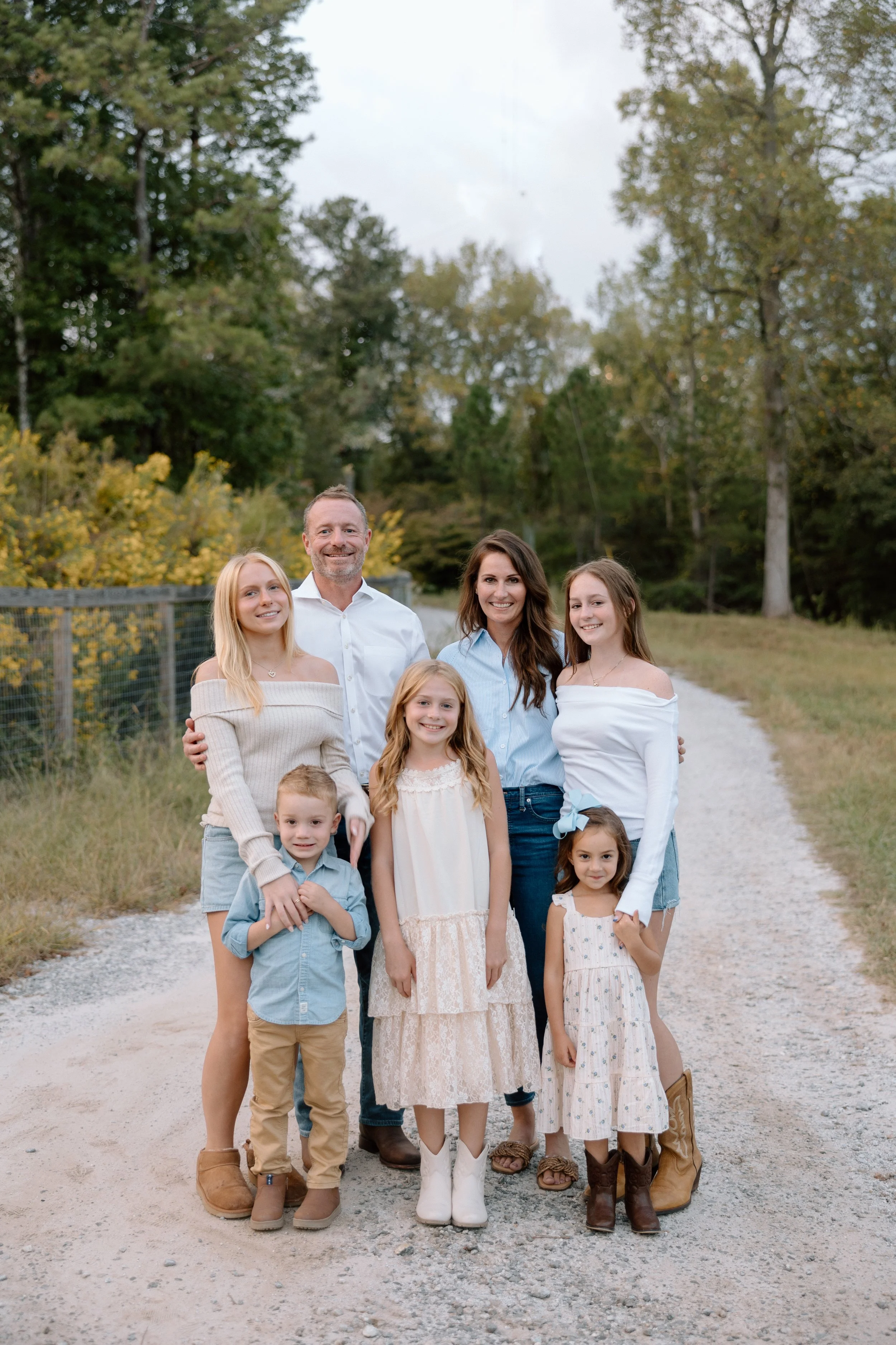 Group of seven people, three adults and four children, standing outdoors on a dirt path with trees in the background. They are smiling at the camera.