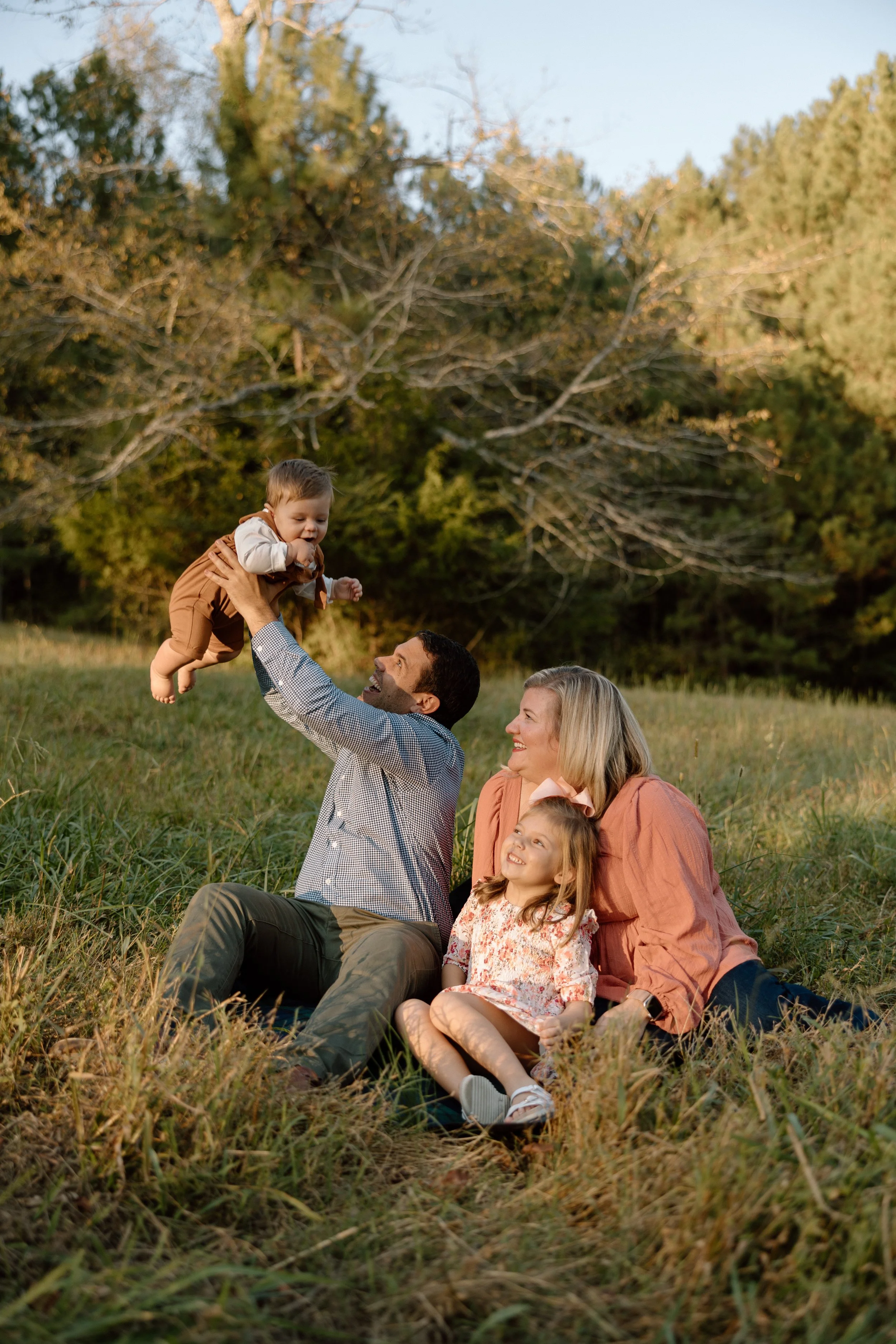 Family of four sitting on grass in a park, with the father throwing a baby in the air, and the mother and daughter watching and smiling, during sunset with trees in the background.