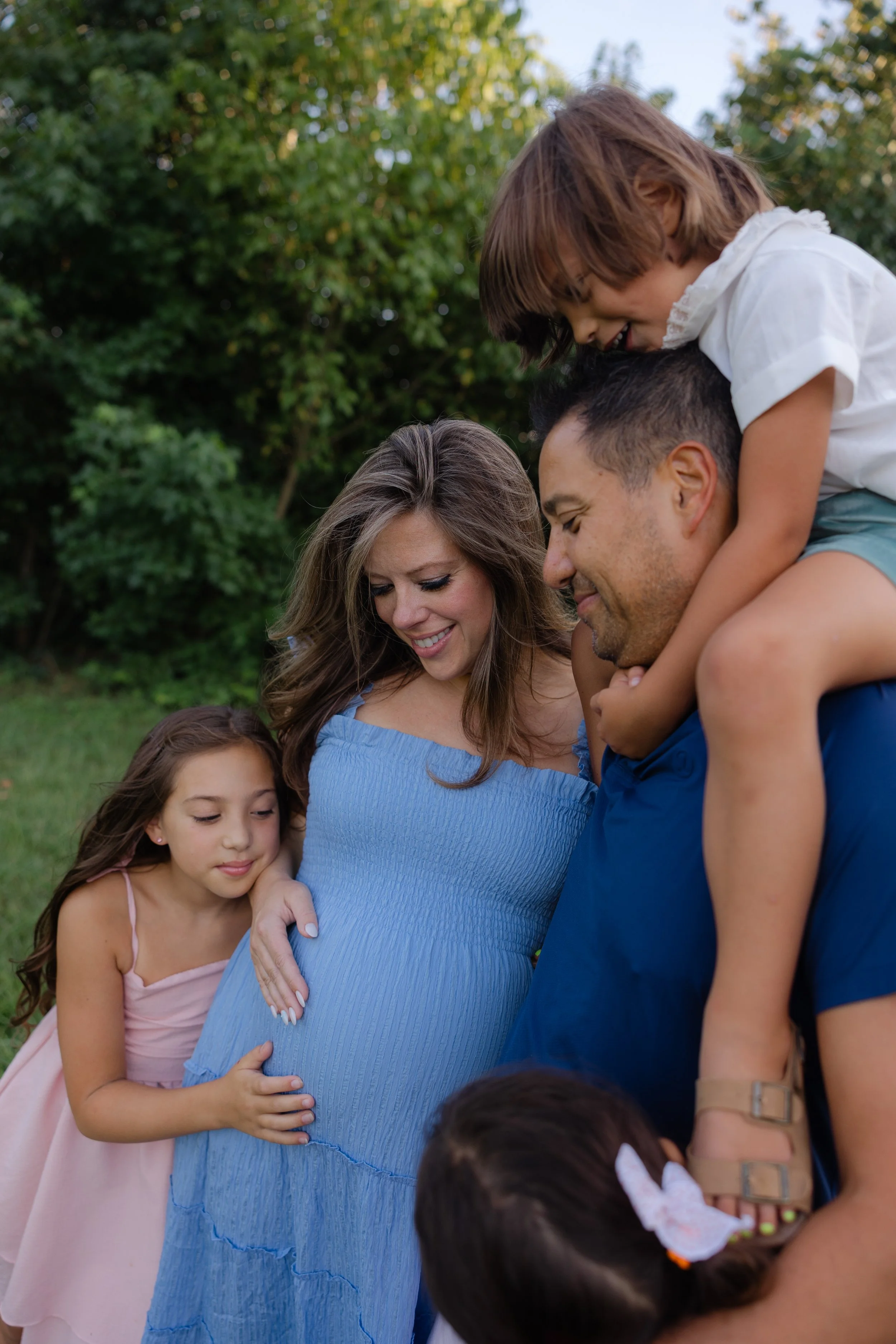 A family of five enjoying a moment outside in a park during the daytime. The pregnant woman is smiling while a man, with a small girl on his shoulders, embraces her. Two young girls are also present, one hugging the pregnant woman and the other standing nearby. The background features green trees and the scene appears joyful and intimate.