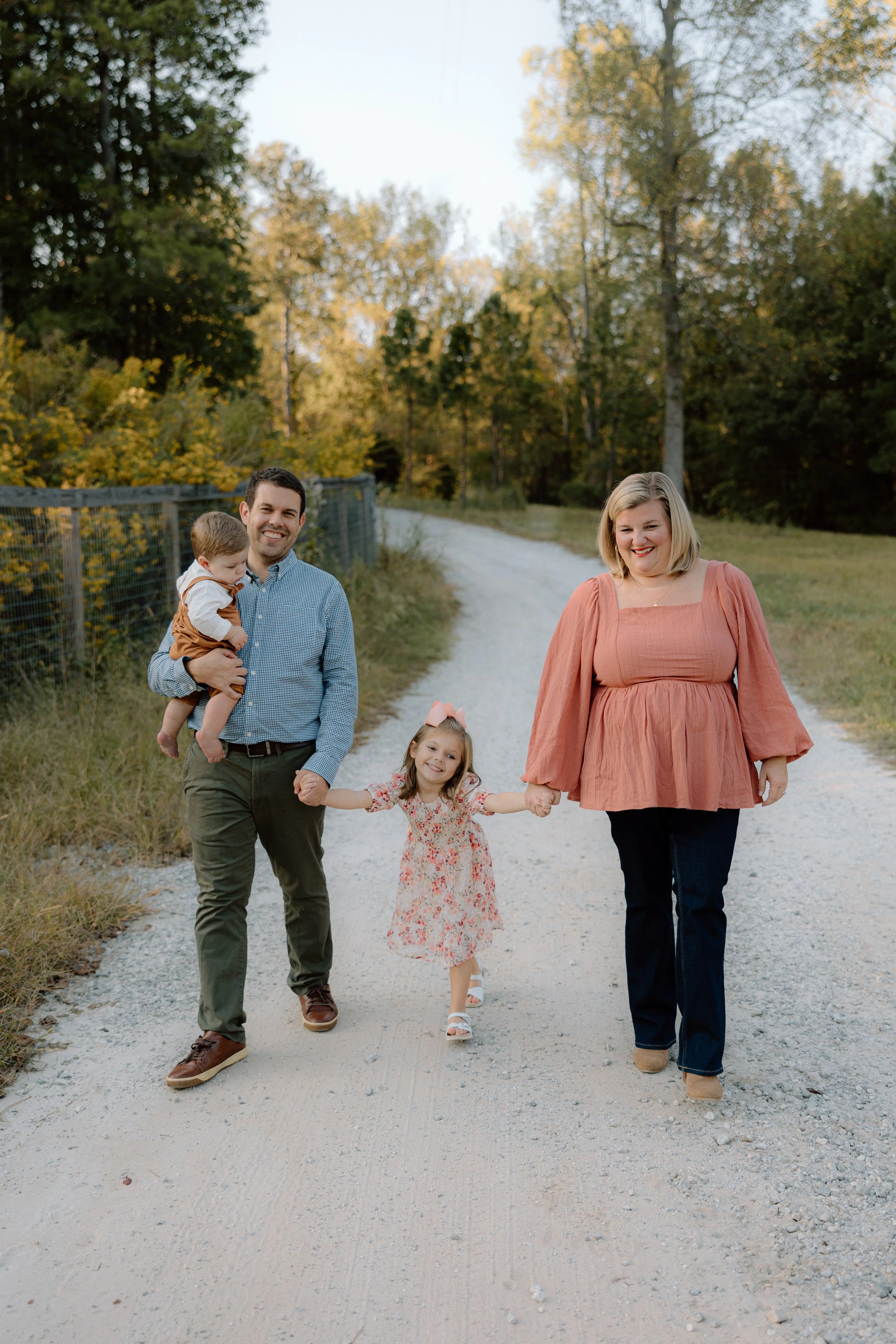 A family of four walking outdoors on a dirt path in autumn, holding hands and smiling. The dad holds a young boy, and the mom walks alongside their daughter, dressed in a floral dress and wearing a pink hair bow.