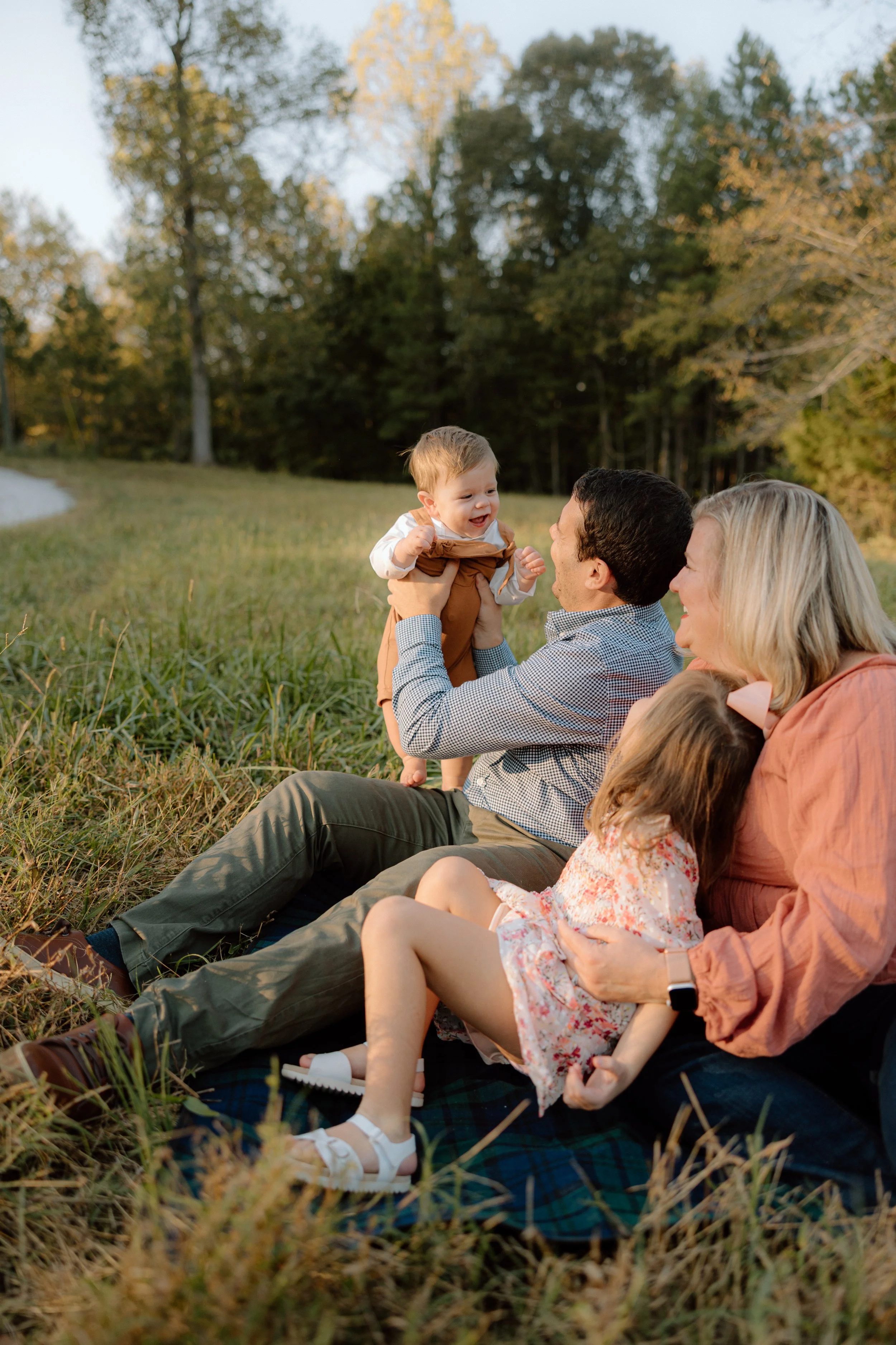 A family enjoying a picnic outdoors in a grassy field surrounded by trees during fall. A man is holding a young boy, and a woman is sitting with a young girl.