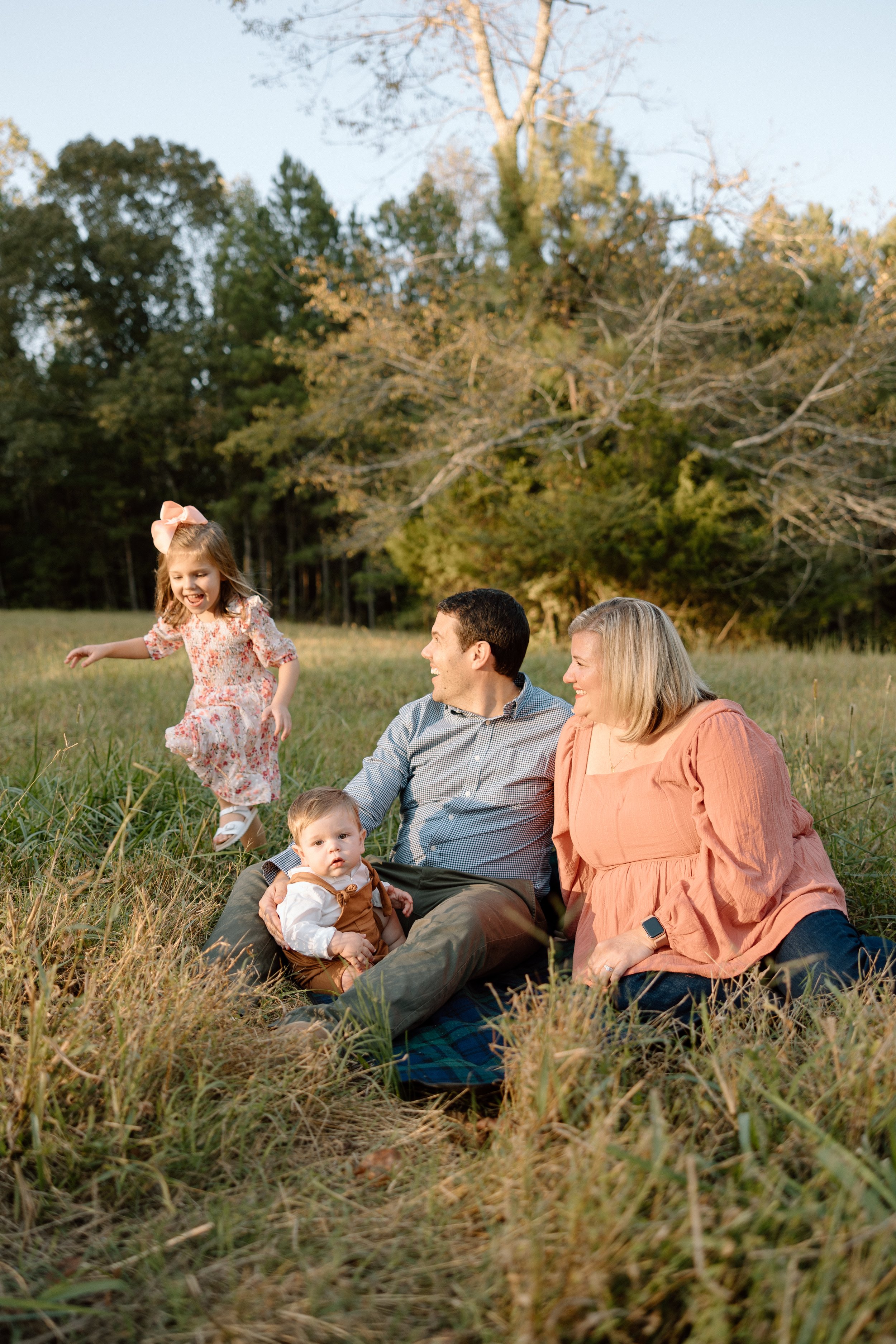 A multigenerational family of four enjoying a day outdoors in a grassy field with trees in the background during late afternoon. The family includes a young girl in a floral dress jumping, an adult man and woman sitting on the grass, and a baby sitti