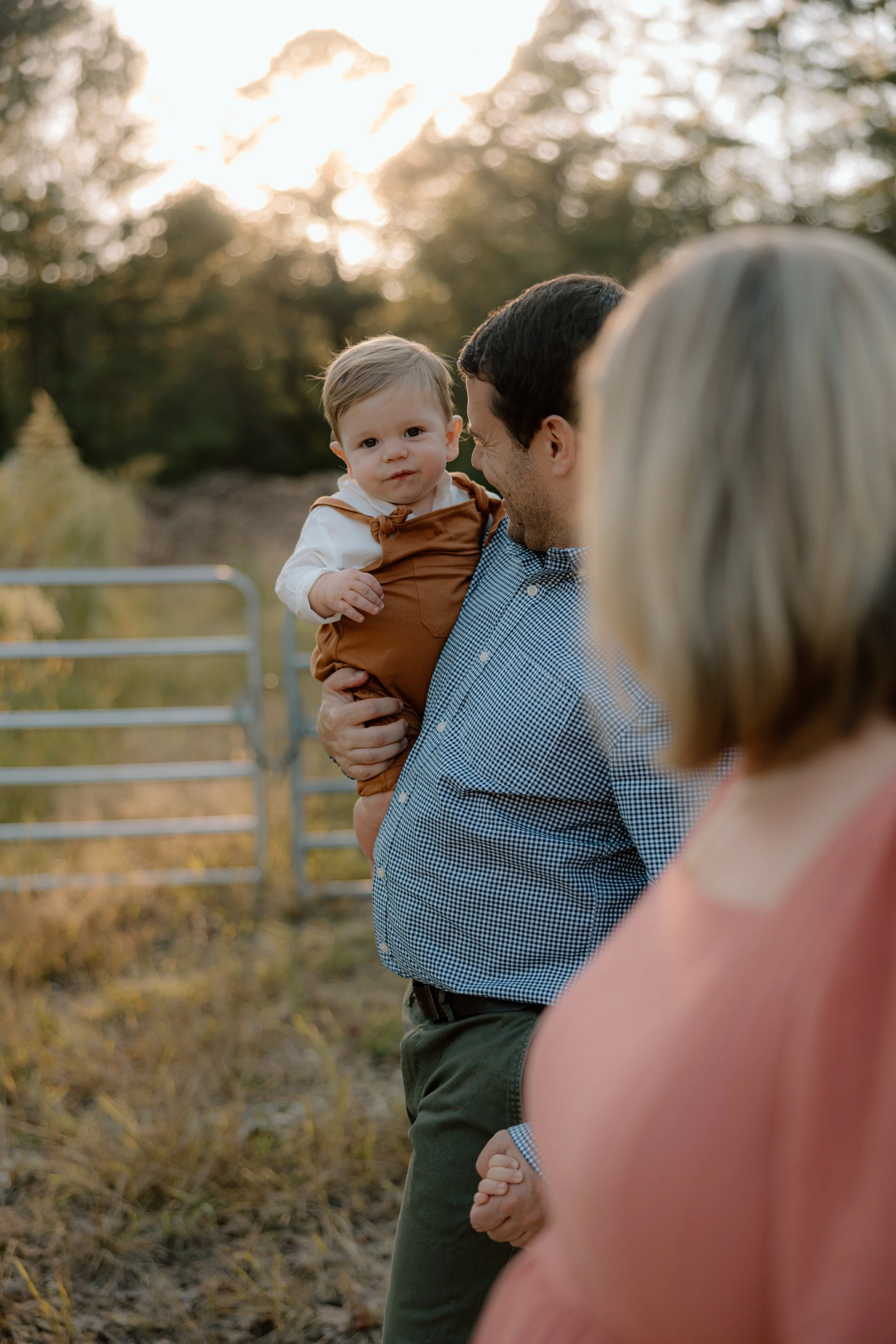 A man holding a young child on his shoulder, with a woman partially visible in the foreground, outdoors during sunset.