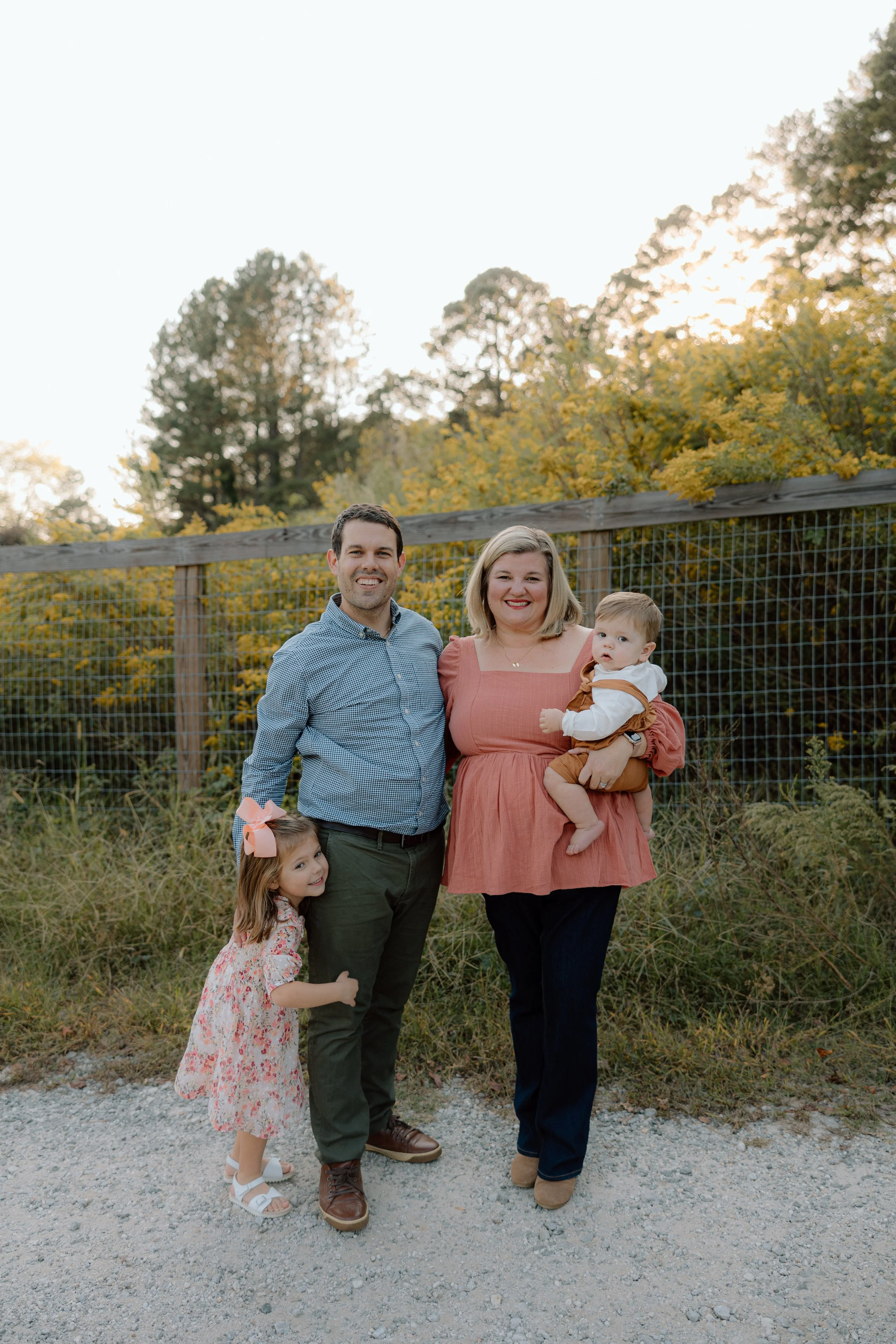 Family of four outdoors, smiling, in front of a wooden fence and trees with yellow leaves, during sunset.