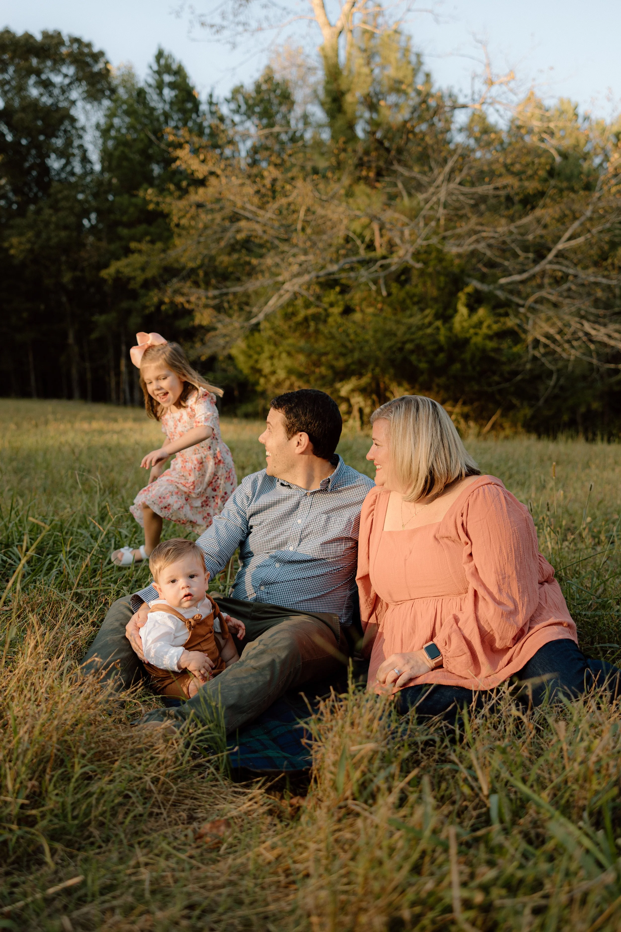 A family of four, including a man, a woman, a young girl, and a baby, sitting on the grass outdoors during sunset, smiling and enjoying each other's company.