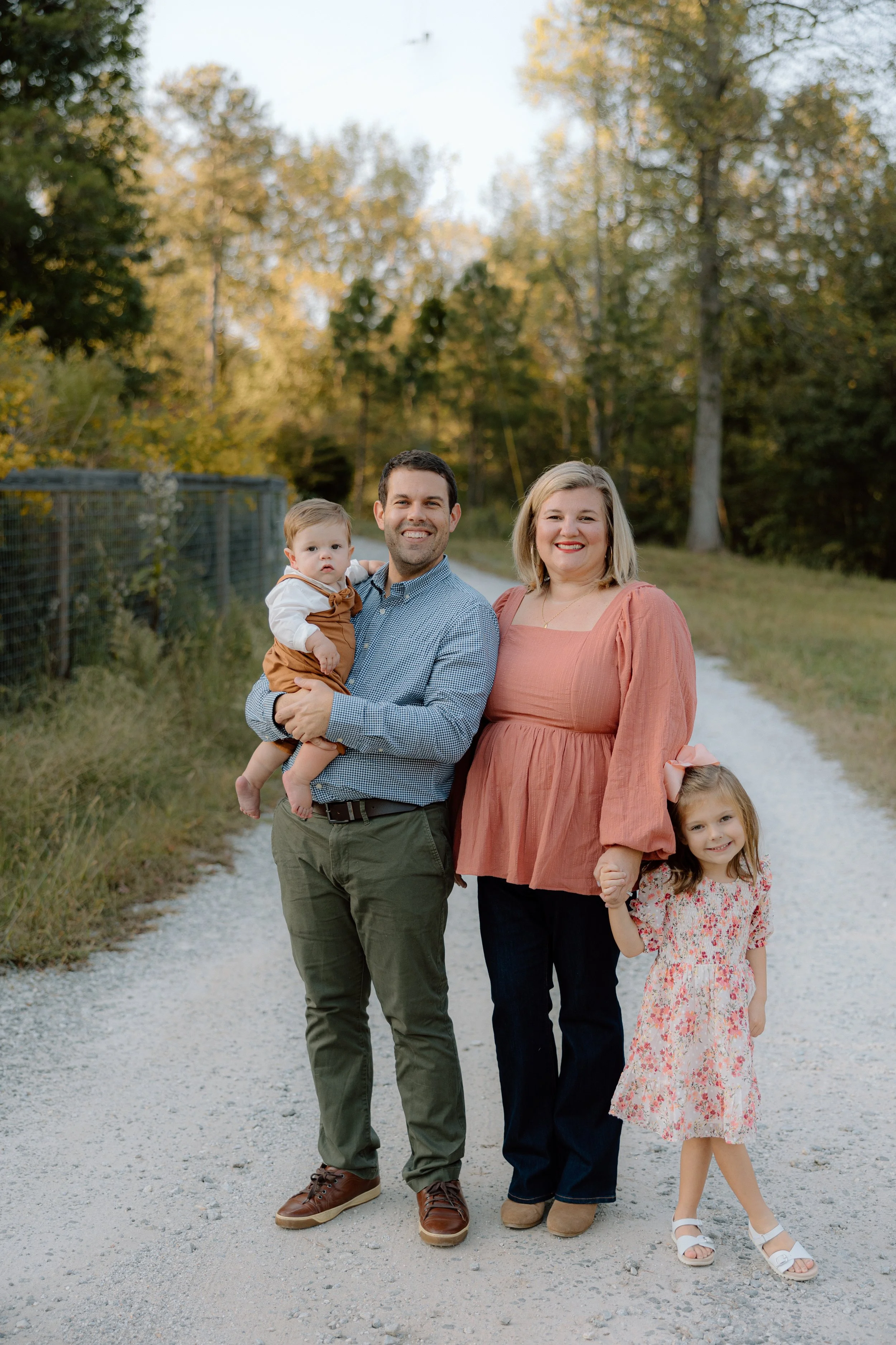 A family of four standing on a gravel path outdoors, holding hands and smiling, with autumn trees in the background.