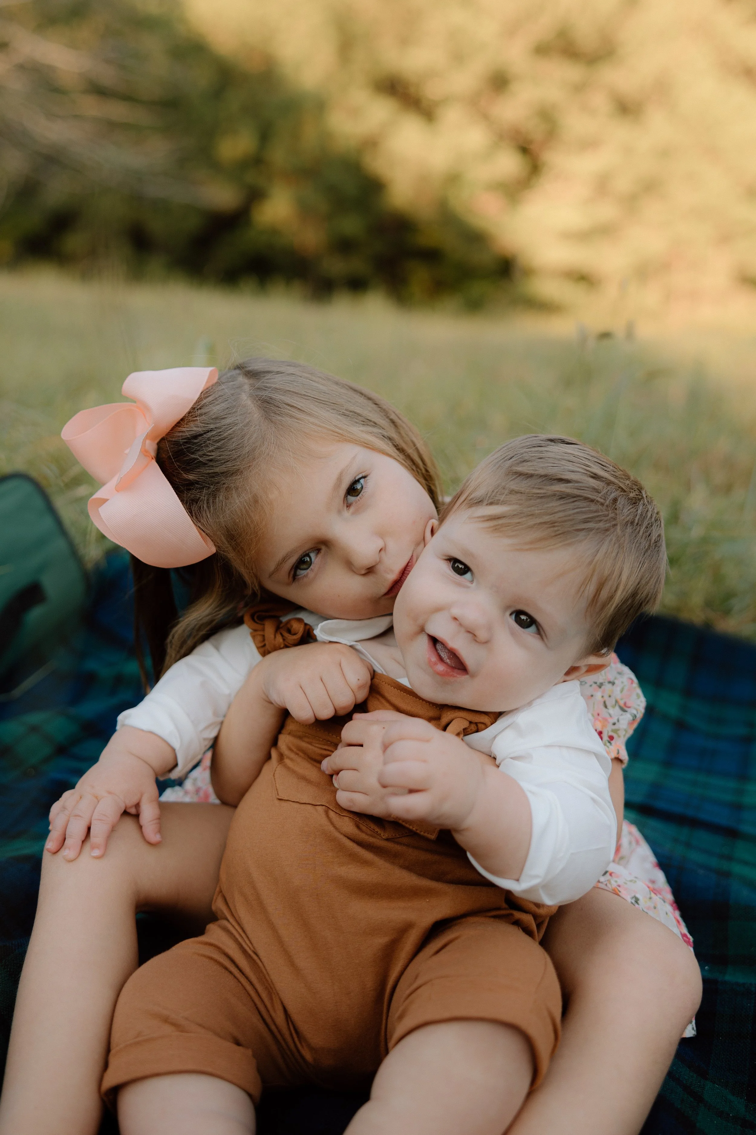 A young girl with a large pink bow in her hair cuddling a baby boy outdoors on a blanket, with green grass and trees in the background.