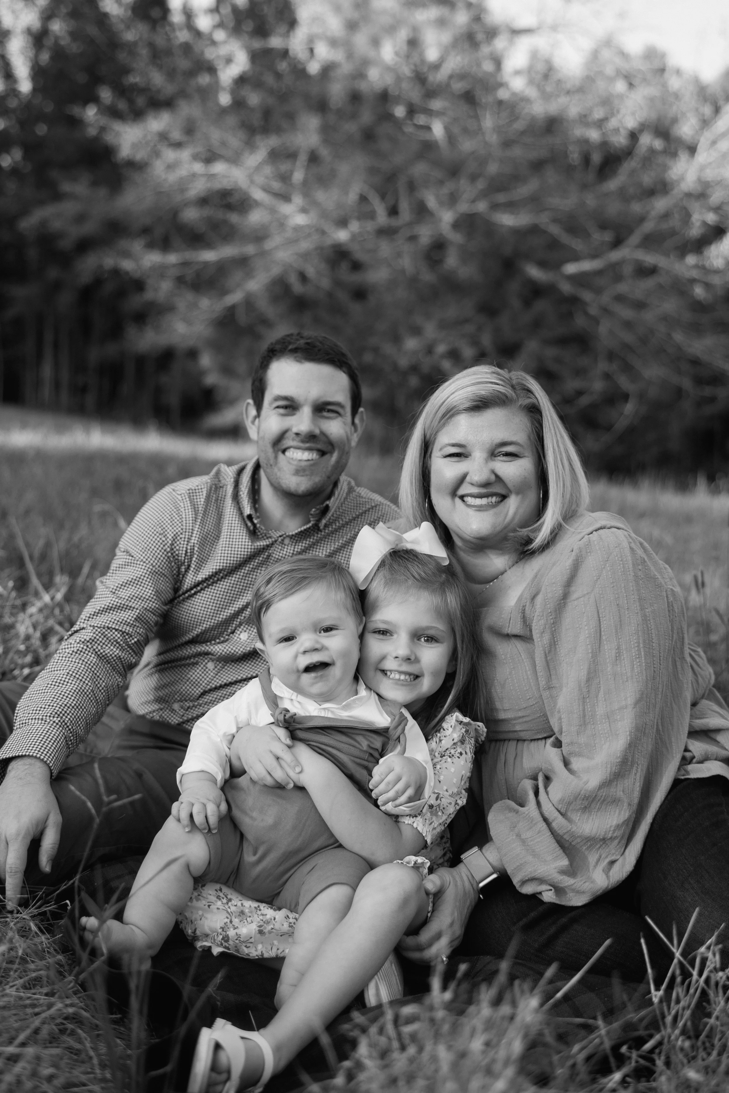 A smiling family of four sitting on grass outdoors, with trees in the background, in a black-and-white photo.