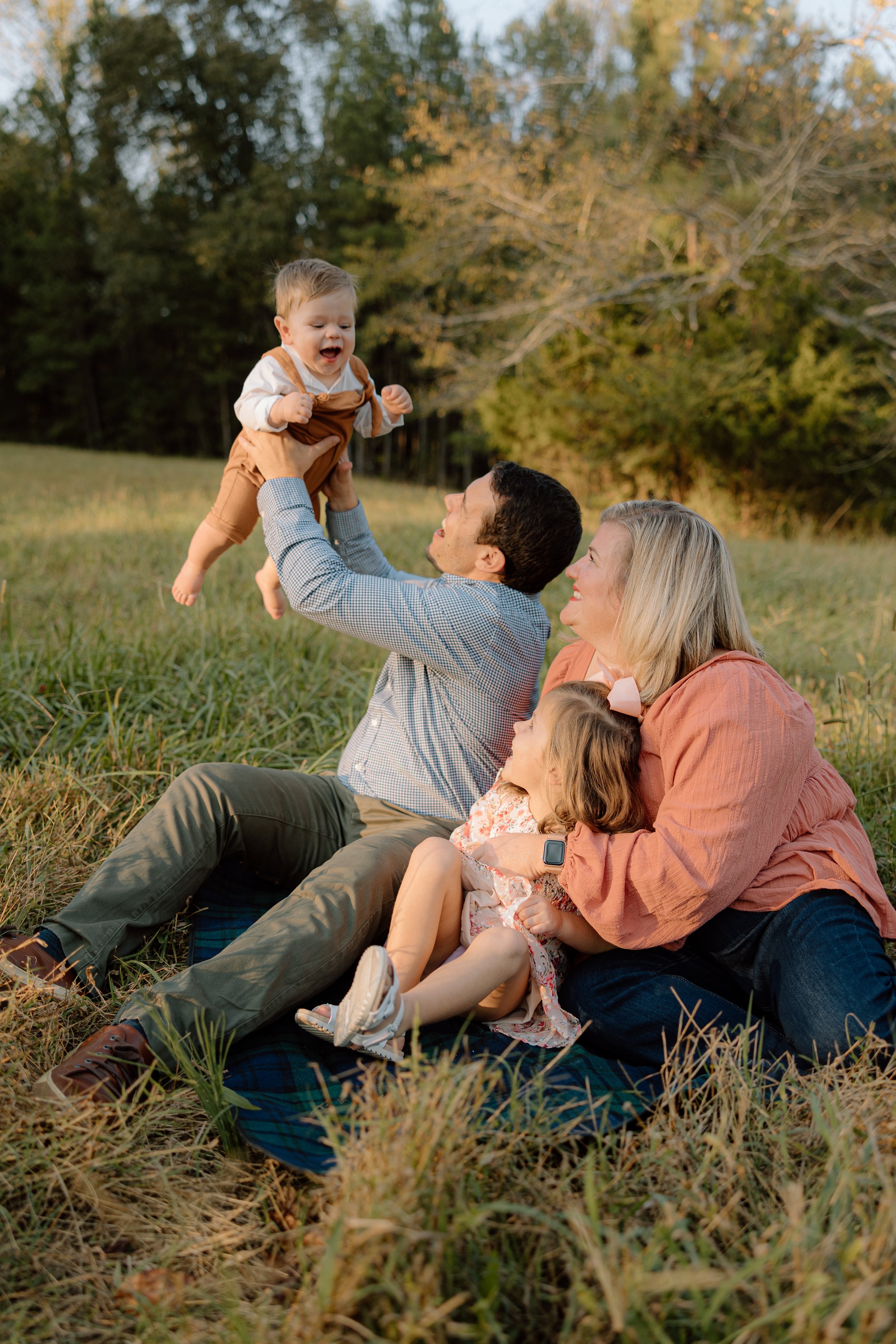 A family of four enjoying a moment outdoors in a grassy field, with a man holding a toddler up in the air, a woman sitting on the grass with a young girl, and autumn trees in the background.
