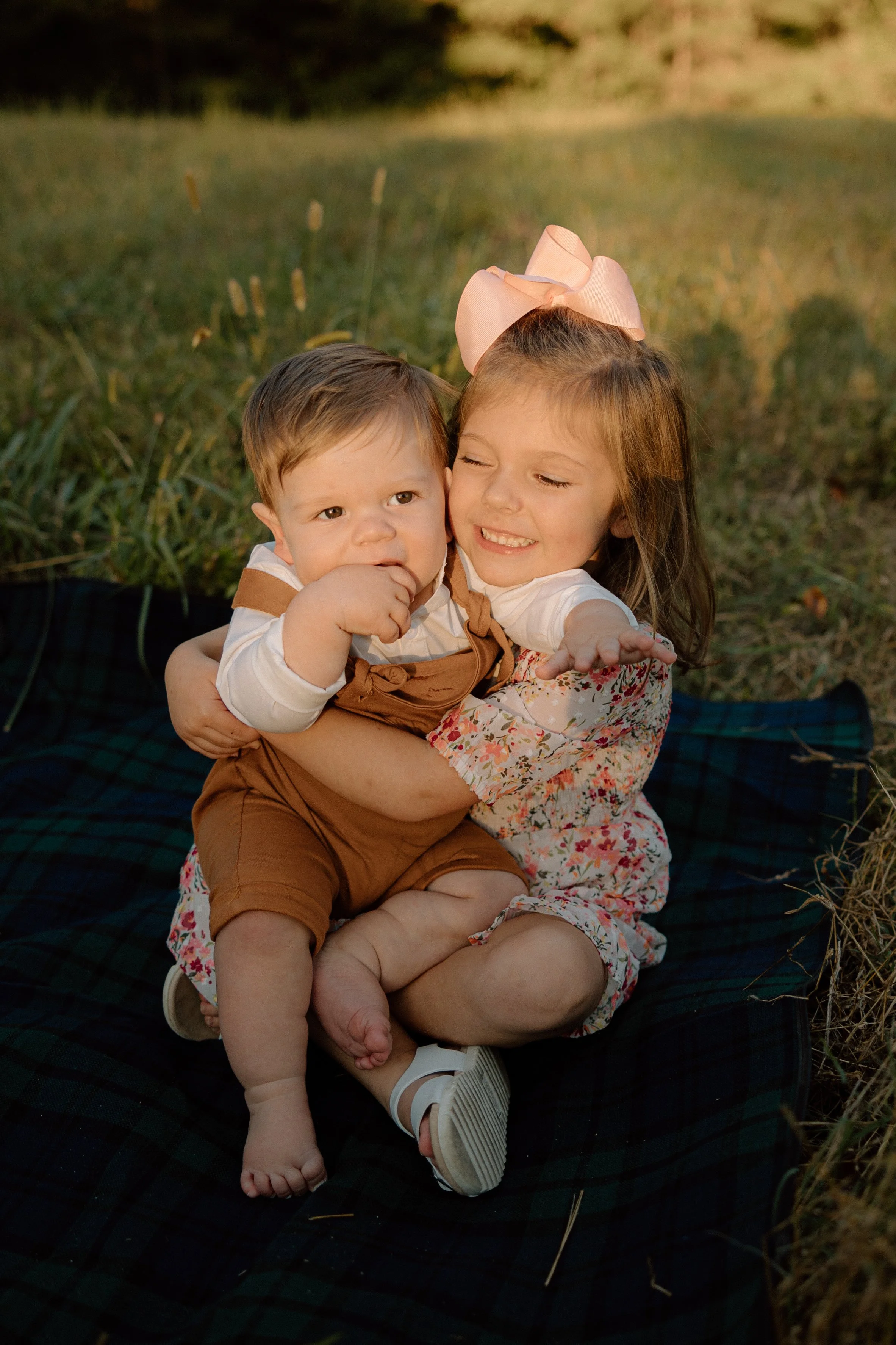 A young girl with a pink bow in her hair hugging a young boy who is sucking his thumb outdoors on a blanket in the grass.