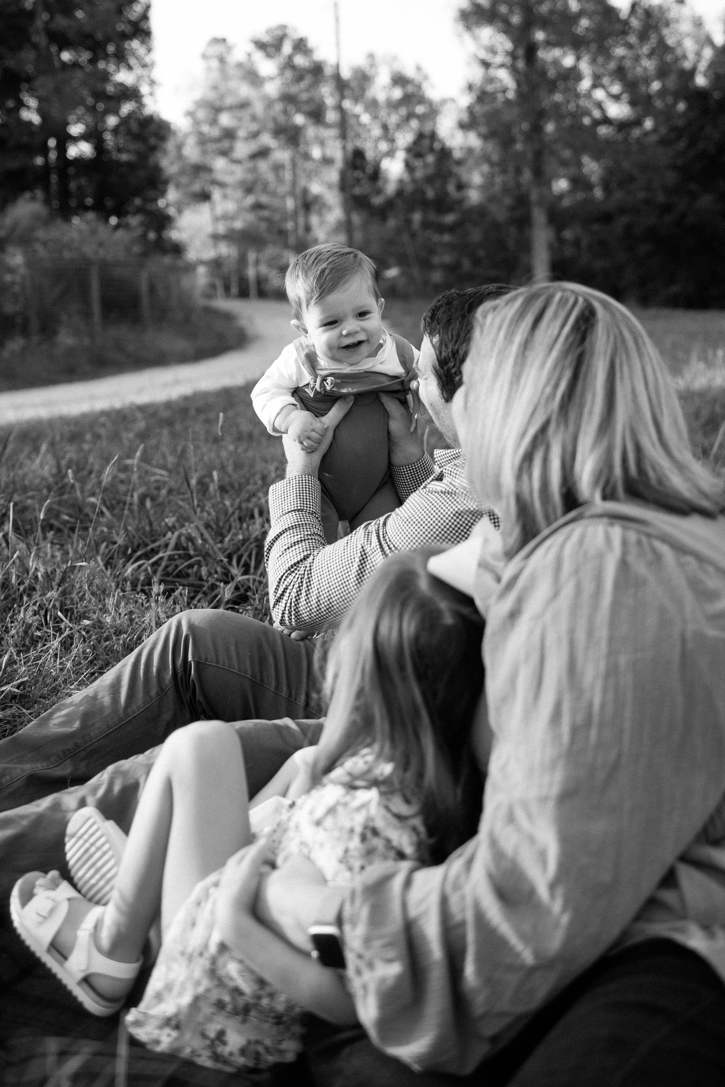 A family, including a baby, a young girl, a man, and a woman, sitting on the grass outdoors. The man is holding a smiling baby, and a young girl is lying next to the woman. Trees and a walking path are in the background.