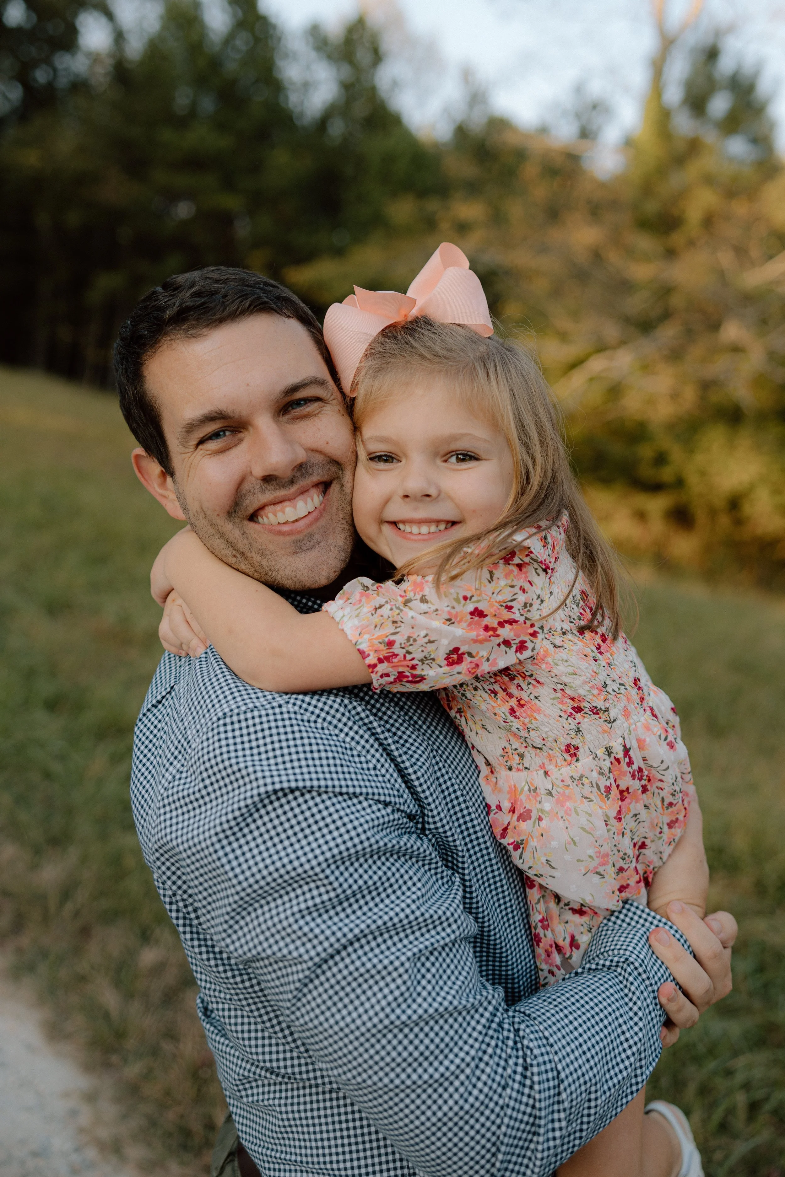 A man holding a young girl in an outdoor setting with trees in the background, both smiling and embracing each other.